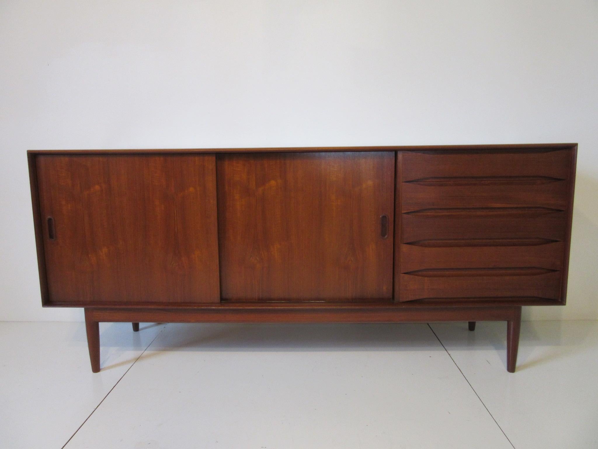 A beautifully crafted teak wood credenza with two bookmatched sliding doors, adjustable shelves and five drawers, two divided with tapering open front areas that serve as pulls. This credenza has a finished backside screwed together with small brass