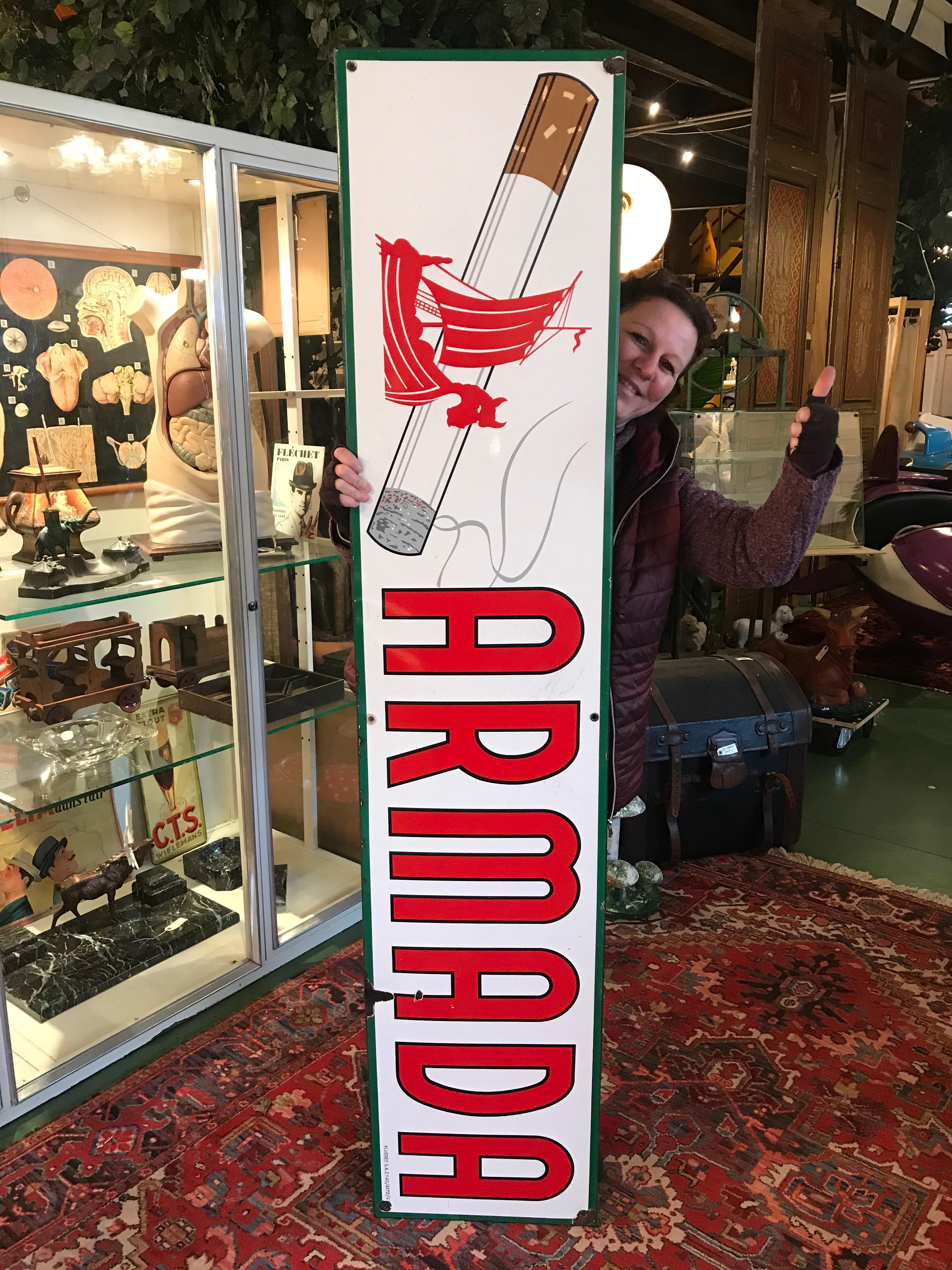 Vintage porcelain sign for Cigarettes Armada.
This cigarette sign is dated 1963. 
This large and long enamel sign dates from the 1960s. It has the color white with the brand name and logo in red and a green border all around. The logo consists of