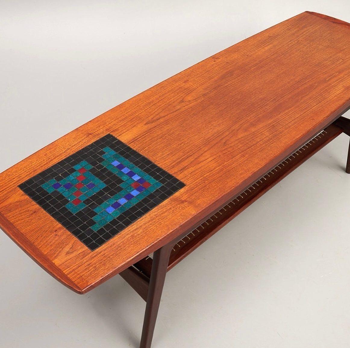 A quintessentially Danish Mid-mod coffee table of solid teak with offset inlaid glass tile mosaic and woven magazine shelf at the underside by Arne Hovmand-Olsen for Mogens Kold. Denmark, 1964.

Measures: 58 inches W x 19 D x 19.