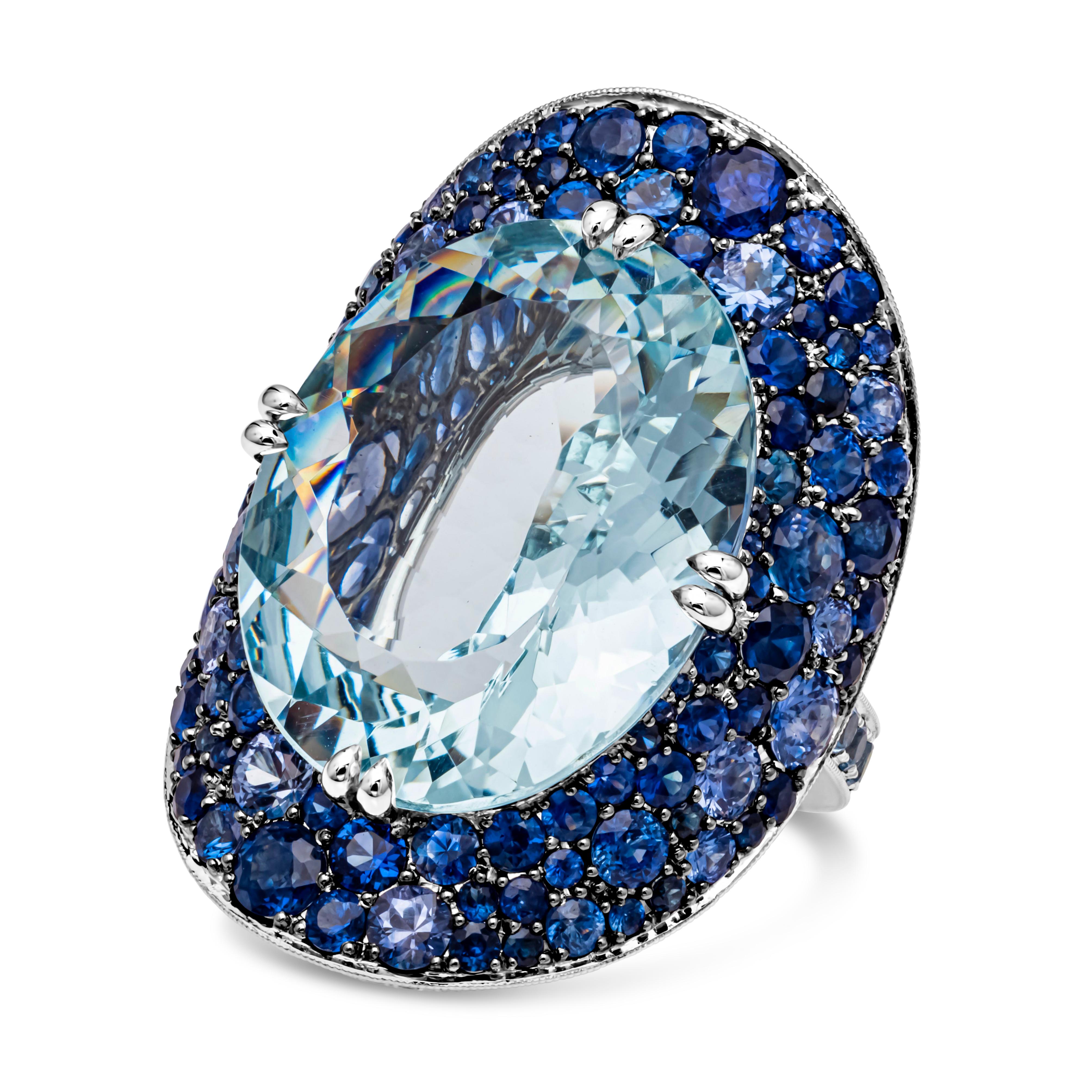 A beautiful and artistic large bonnet ring, showcasing an oval cut aquamarine weighing 19.69 carats total, set on a timeless eight prong basket setting made of 18K white gold. Surrounded by round brilliant blue sapphires and aquamarines in a pavé