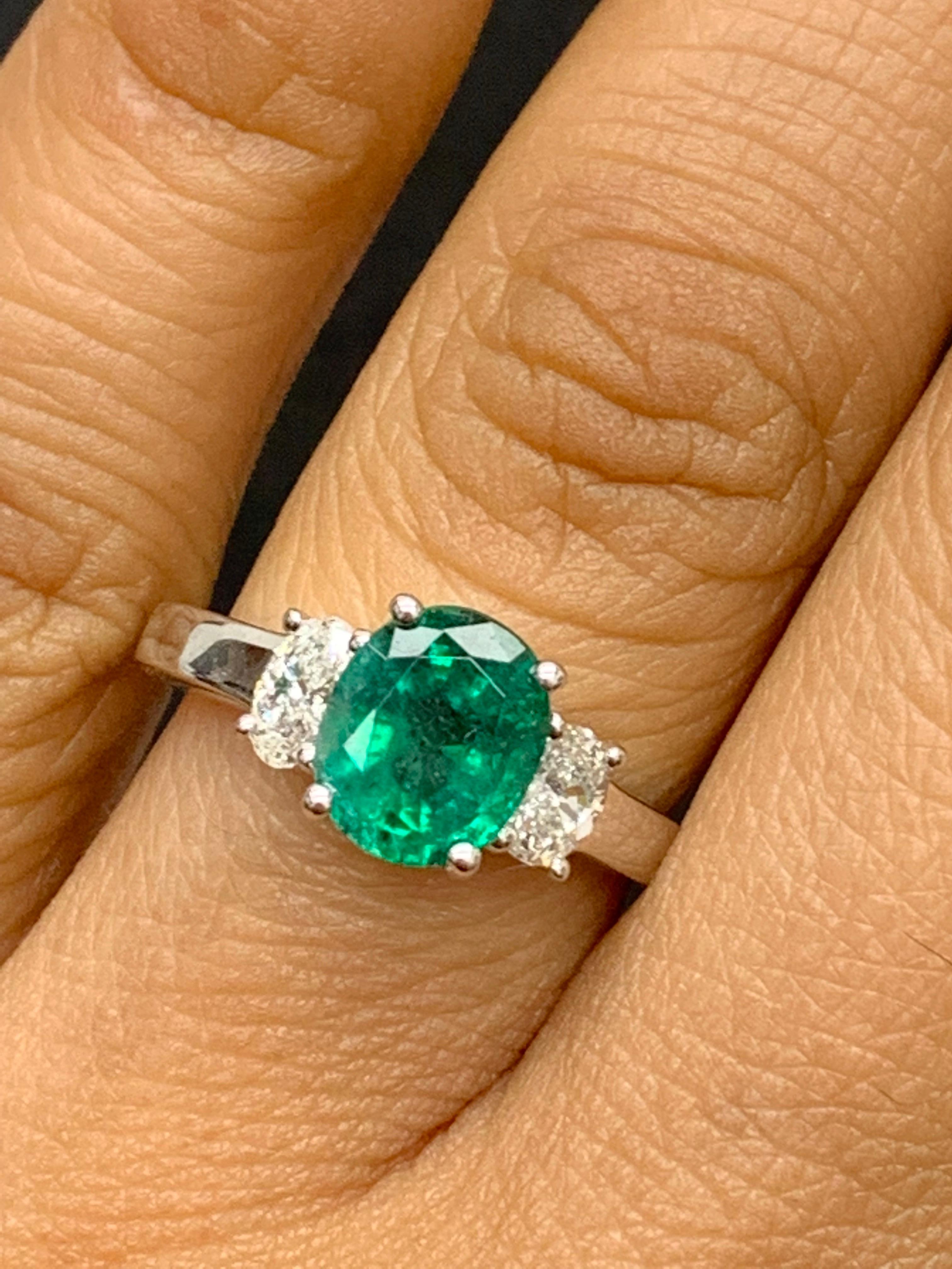 Showcases an Oval cut, Vibrant color green Emerald weighing 1.97 carats, flanked by two brilliant oval cut diamonds weighing 0.48 carats. Elegantly set in 18k White Gold.