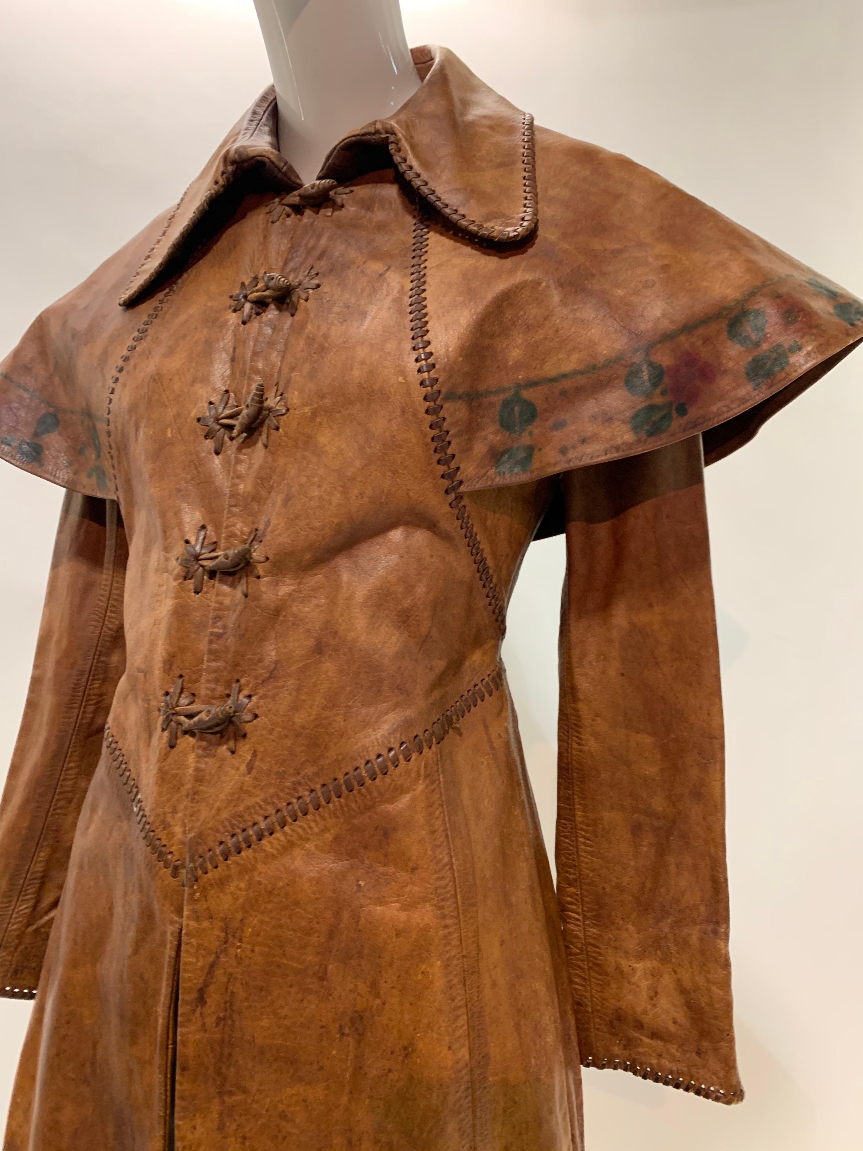 A very unusual 1970s hand-made distressed leather women's trench coat in a fairytale style with integral caplet, toggle front closures and hand-painted floral trim. Pointed Elizabethan waistline at front and back. Size 4-6. 