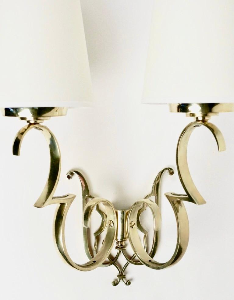 Composed of two arms in gilded brass forming curves and ascending half-curves, dressed with conical shades of unbleached cotton.
The base of the wall lamp is made of two scalloped rods in gilded brass.

2 bulbs per sconce.