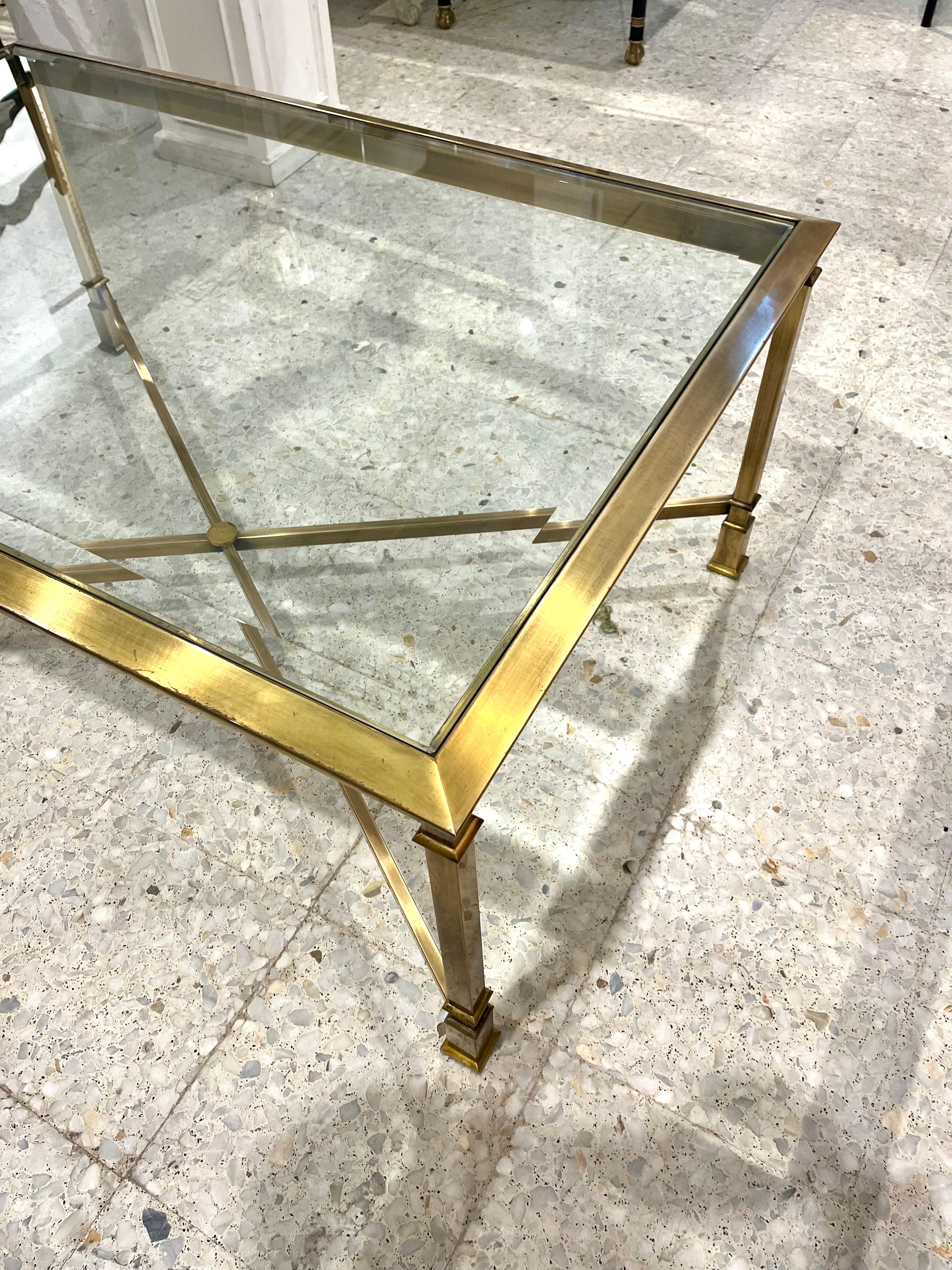 Table basse ou table de cocktail espagnole des années 70, en laiton massif et plateau en verre biseauté, avec un croisillon en forme de X à la base. Il repose sur quatre pieds à décor géométrique carré.