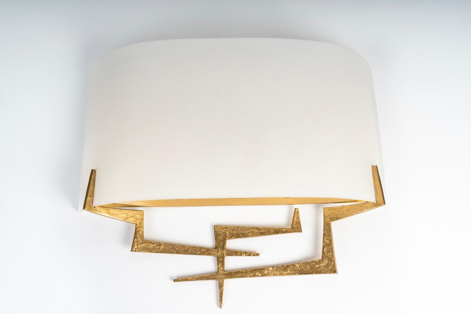 Composta da una staffa da parete in ferro battuto martellato con una patina dorata, decorata nella parte inferiore con un attraente motivo grafico che corre per tutta la lunghezza dell'applique e nella parte superiore con un riflettore in lamiera