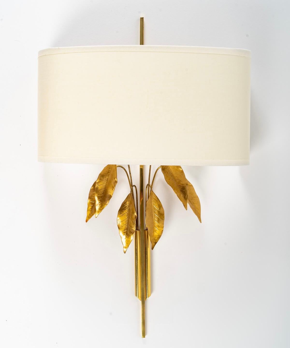 Composed of a long square section stem in gilded brass decorated with two small tubes on either side of the stem in which are placed fine stems of gilded brass foliage.
On the upper part, the wall lamp is dressed with a shade in off-white cotton