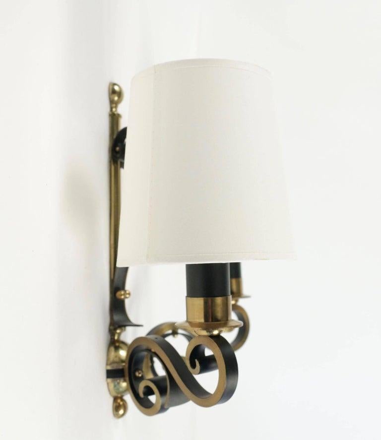 The base of the wall lamp is made of a vertical rod in gilded brass underlined by two black wrought iron volutes.
In the lower part, a gilded brass ball supports the two arms of light in the form of gilded brass volutes, they are underlined by