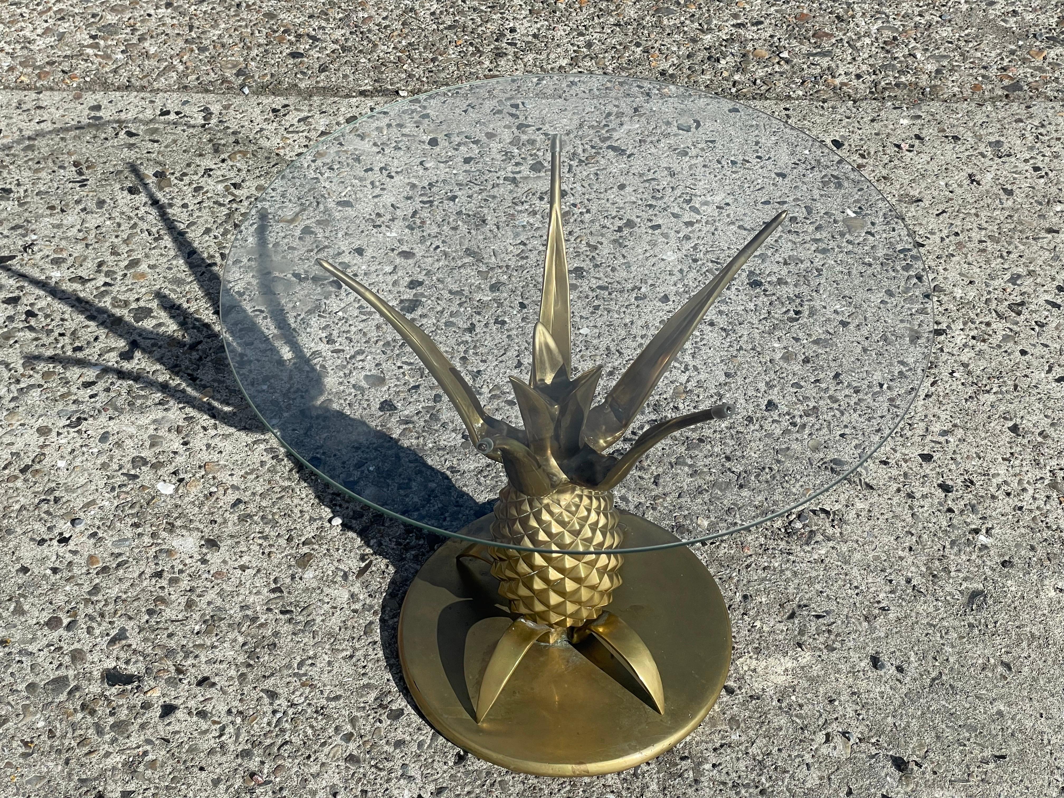 The pineapple side table in brass with glass is a glamorous Hollywood Regency style piece from the 1970s that is sure to make a statement in any room. The table features a stunning brass pineapple base with intricate detailing and a sleek glass top