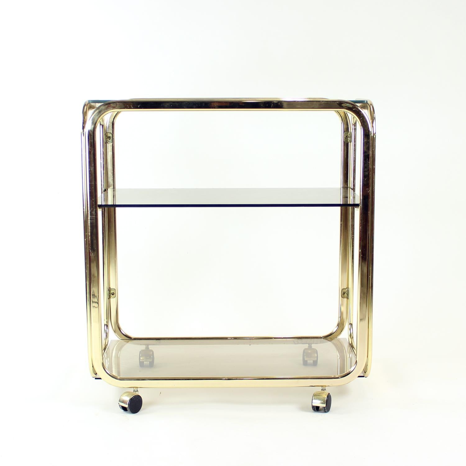 Superbe trouvaille vintage ! Il s'agit d'un open bar des années 1970 sur quatre roues. La construction est en métal avec une finition dorée d'origine. Le bar comporte trois étagères en verre fumé. Le verre est en excellent état et ne présente aucun