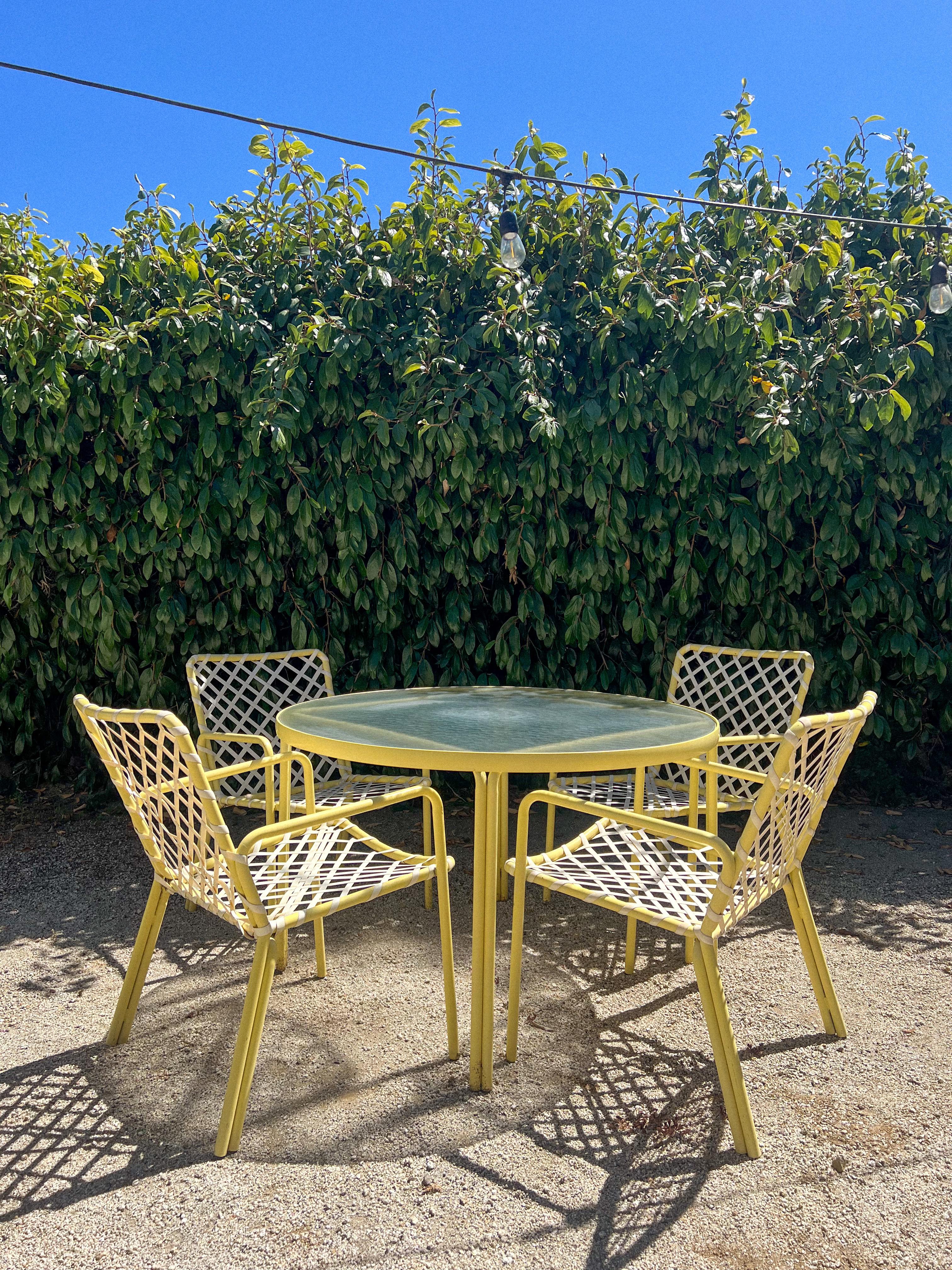 A timeless vintage patio set by Brown Jordan, featuring their iconic Tamiami collection design. This set includes a round glass-top table and four armchairs with powder-coated aluminum frames in a sunny yellow finish. The chairs showcase the