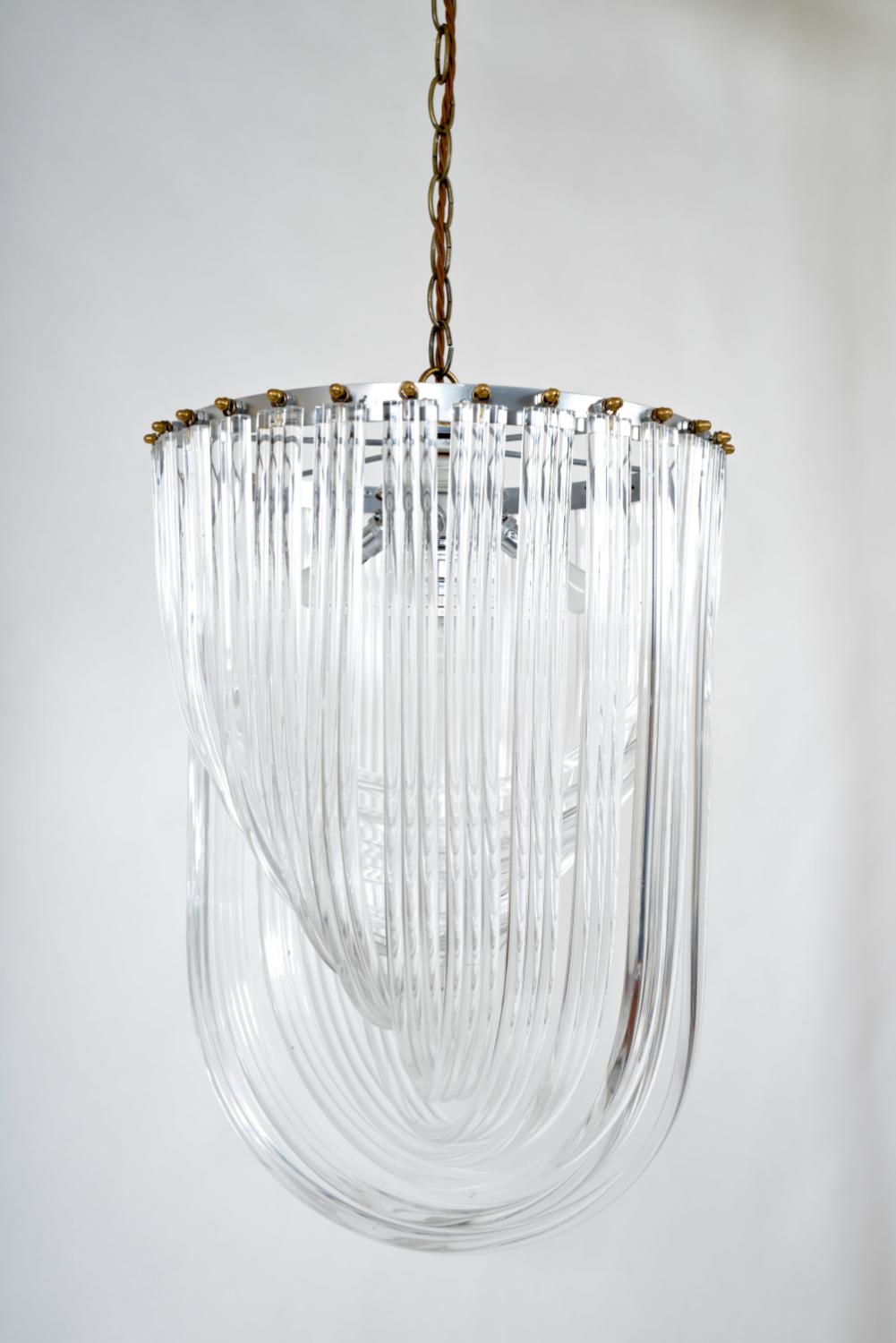 This stunning 1970s American Lucite ribbon and brass chandelier comprises of U-shaped thick bands of cascading Lucite in different lengths that are suspended from a chrome frame with brass fixings. The large Lucite is illuminated by four bulbs at