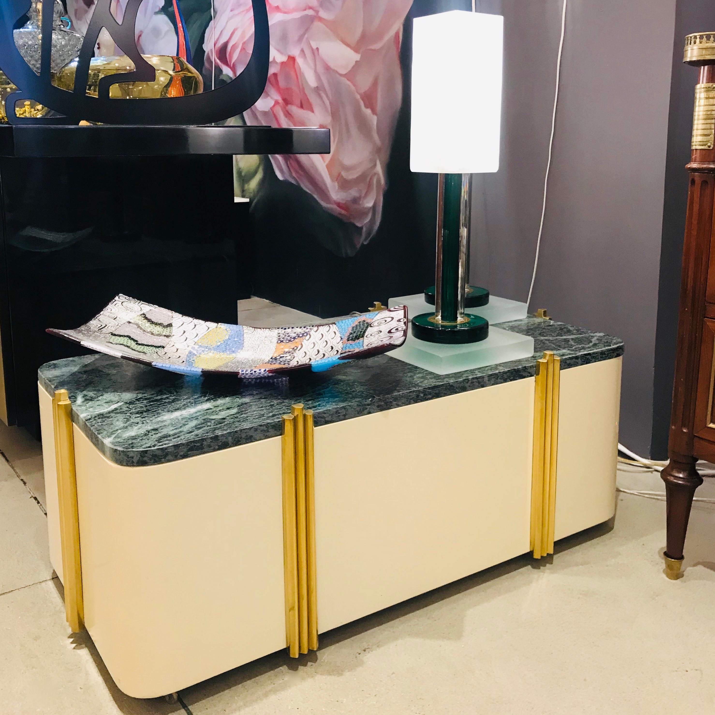 A very interesting and rare Italian modern sofa table on brass casters, of Art Deco design, entirely handcrafted in ivory white lacquered wood topped with green Italian Verde Issorie marble, elegantly decorated on each side with vertical brass