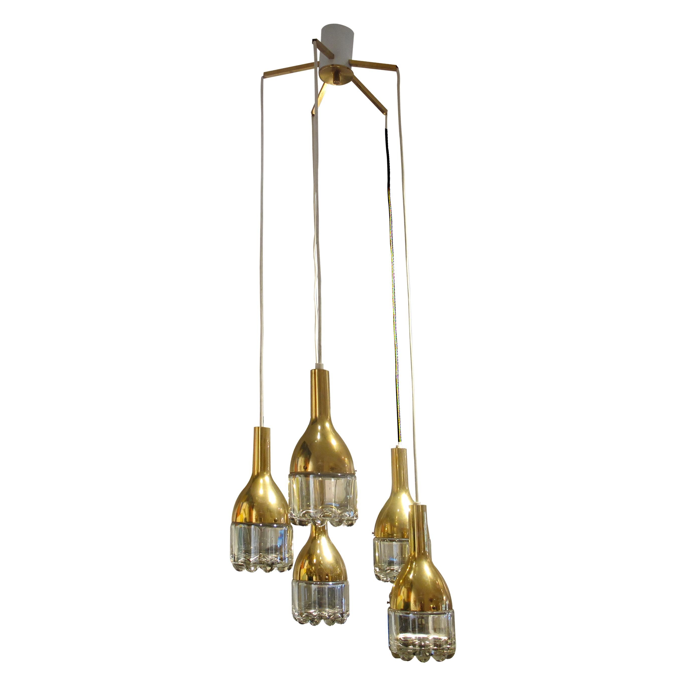 This Belgian chandelier from the 1970s features a cascade design with five pendant lights suspended at varying heights. Each pendant has a brass-finished metal body and a clear moulded glass diffuser at the base. The glass has a rounded, textured