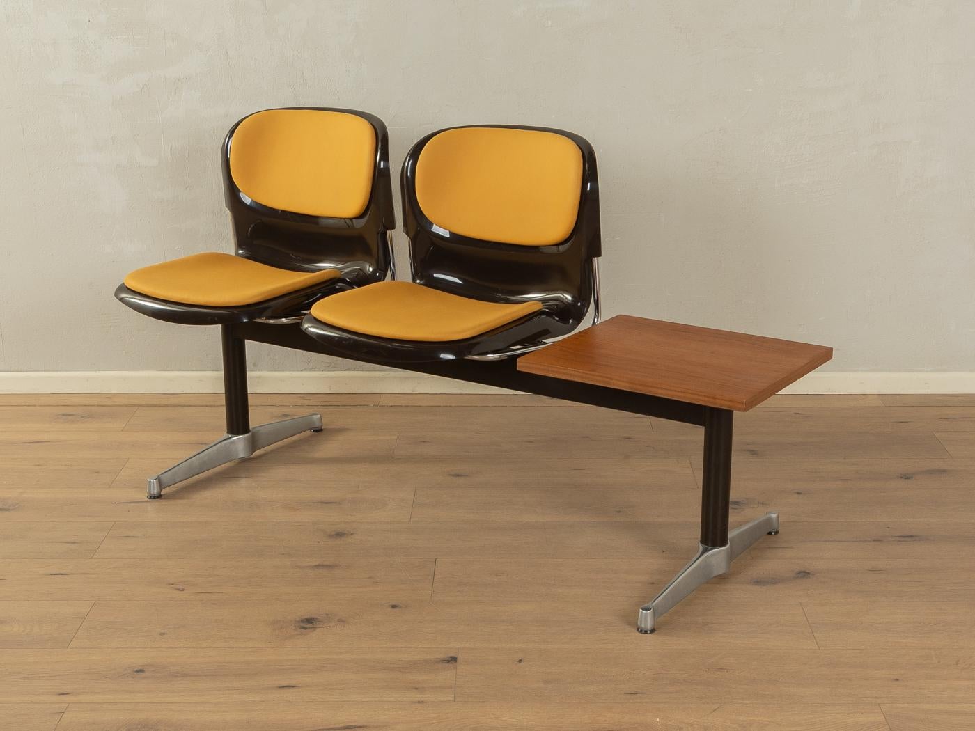 Classic waiting bench from the 1970s. High-quality steel and stainless steel frame with two plastic seat shells in brown and a table top in teak veneer. The seat shells have been reupholstered and covered with a high-quality leatherette in ochre.