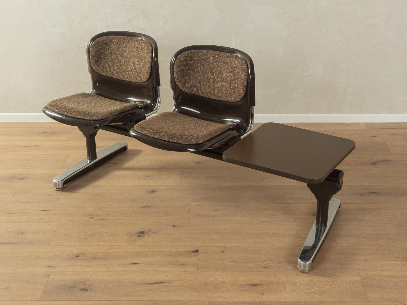 Banco de espera clásico de los años 70. Estructura de acero y acero inoxidable de alta calidad con dos carcasas de asiento de plástico en marrón y tablero de mesa con revestimiento HPL original y borde de plástico en marrón. El tejido original se ha