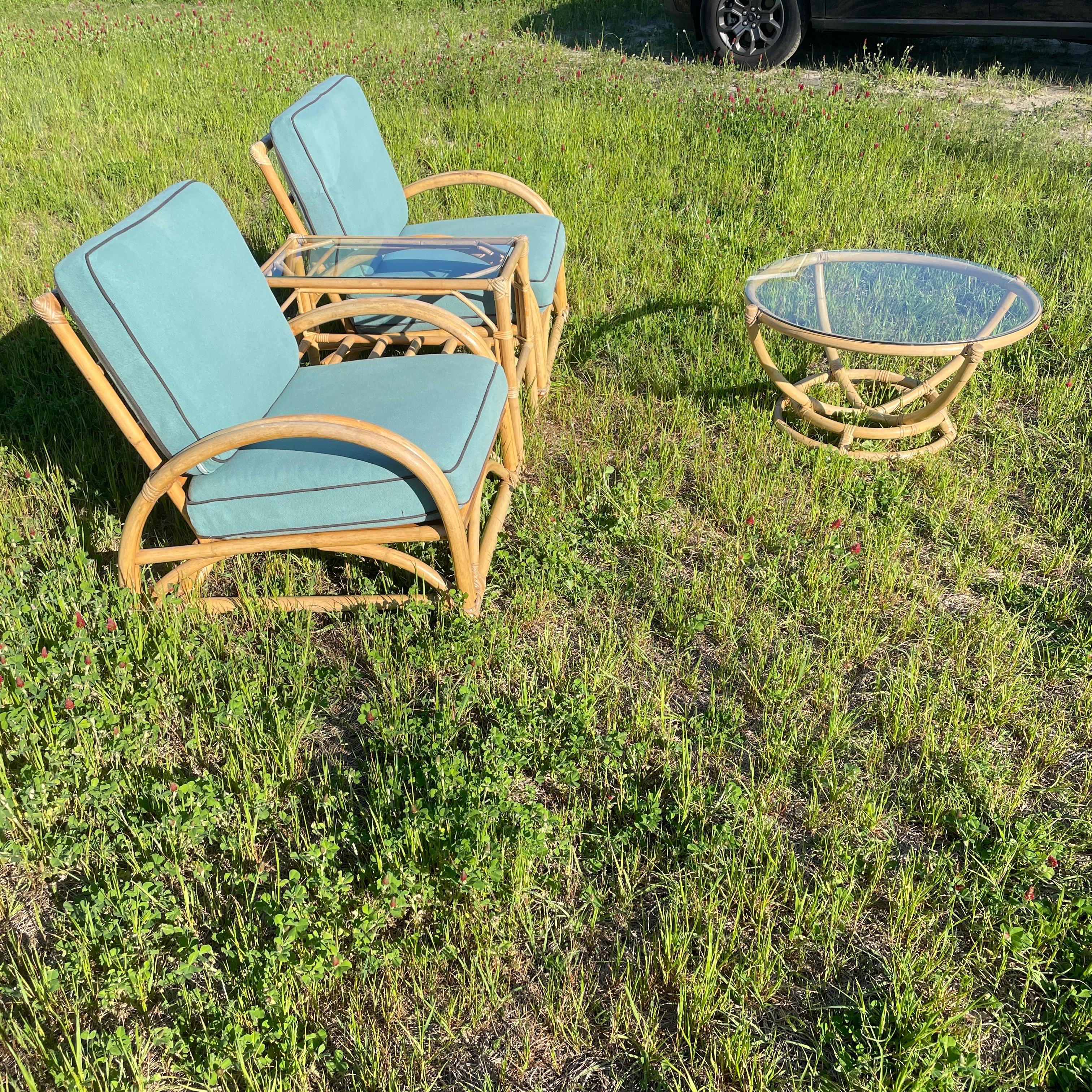 Paul Frankl style four piece rattan patio furniture set, which includes two comfortable arm chairs and two glass top tables. One table being a round glass top coffee table and the second being a glass top side table with a shelf underneath. All of
