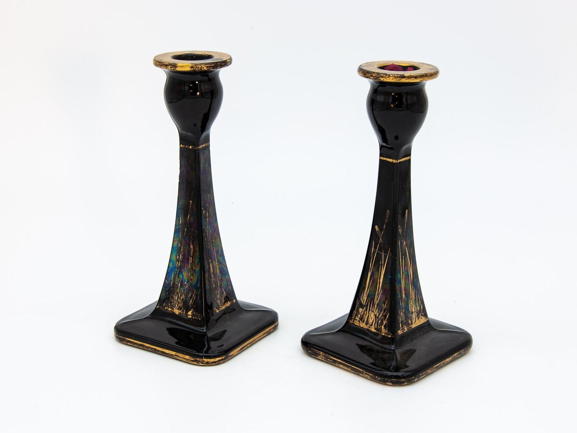 A pair of 1970s era black & gold candle holders with a carnival glass finish. Each candlestick has a tapered square column terminating on a square base. The cup is rounded with a gilt finish. The black finish column has a painted gold design on all