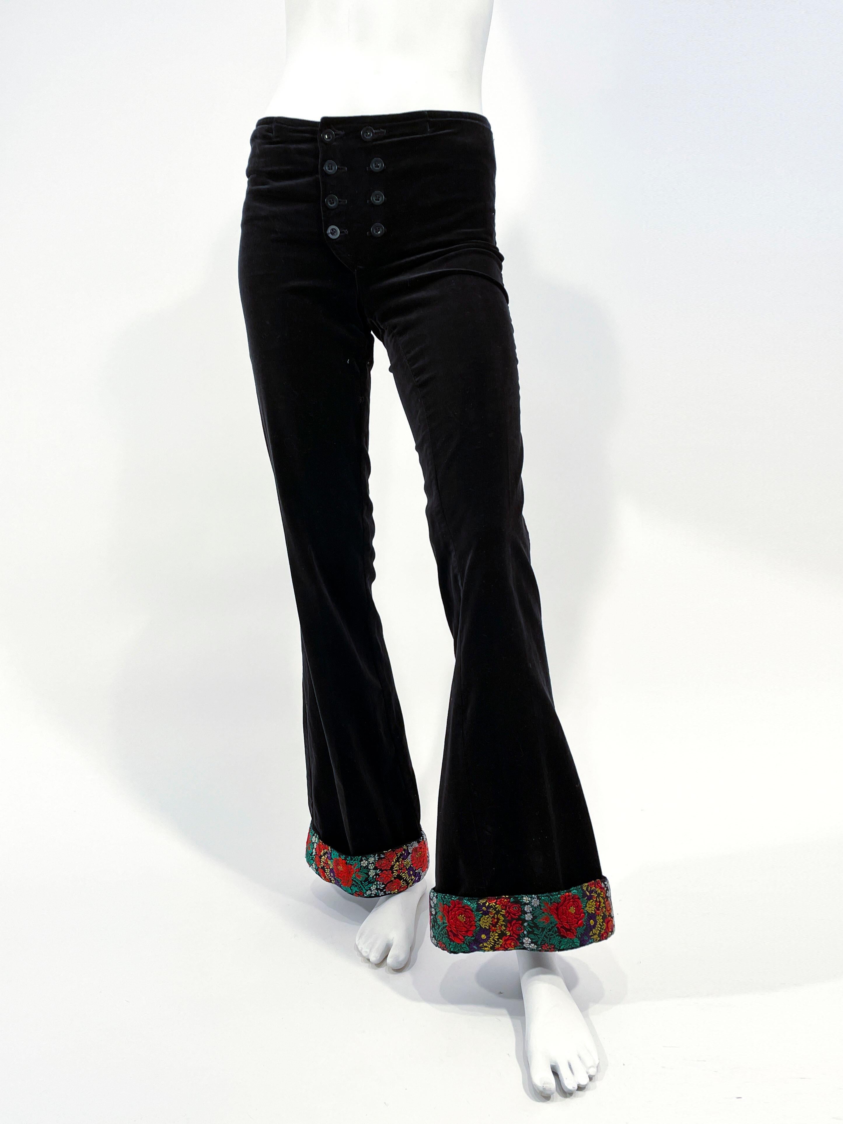 1970s custom made black velvet bellbottom pants with a double button fly on its low-rise hip hugger waist. The wide rolled cuffs on the bellbottom are finished with a vibrant multicolored floral trim. These pants are unlined with two pockets on the