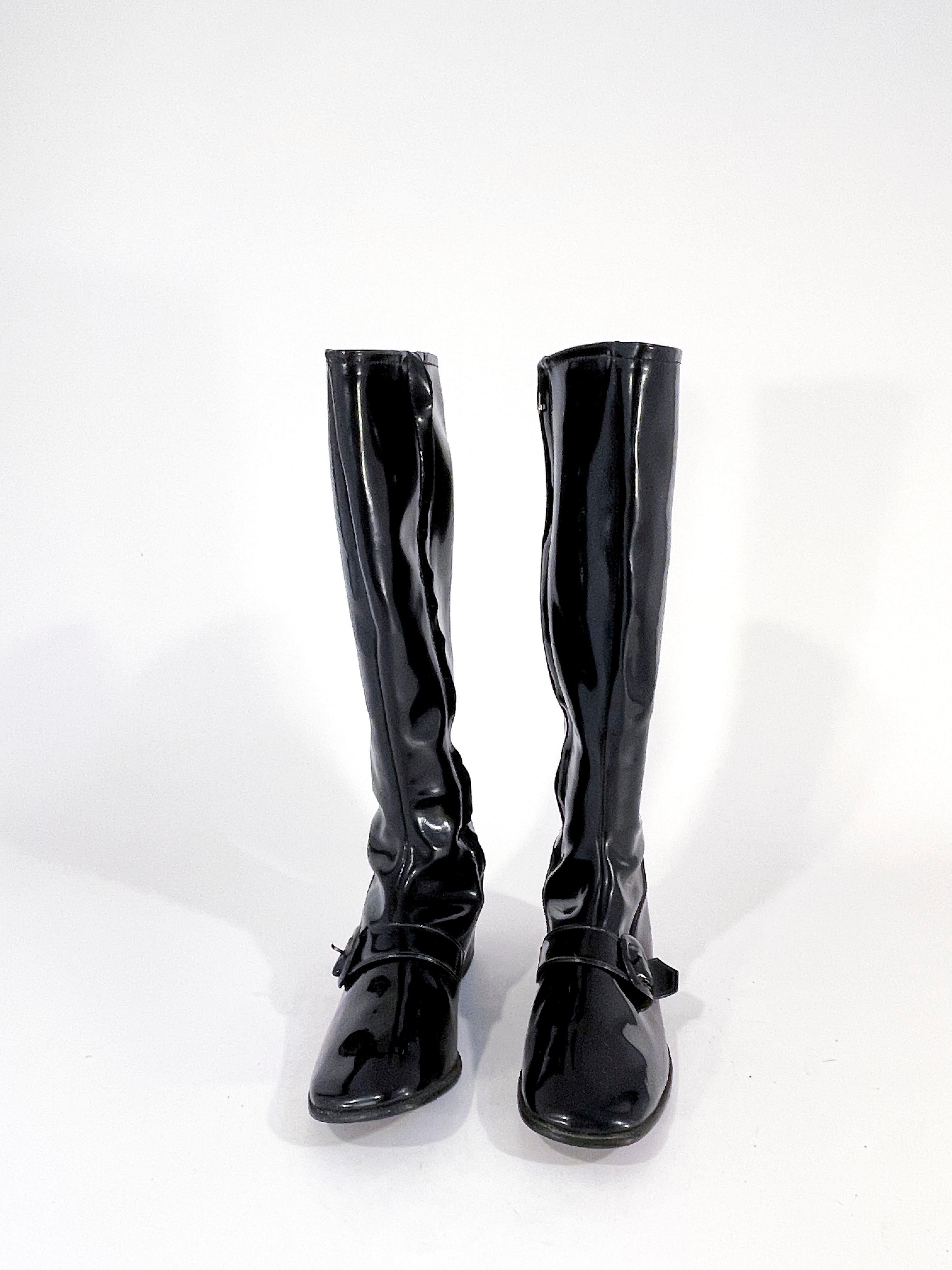 1970s shiny black vinyl knee-high go go boots with decorative strap and buckle over the bridge of the foot. The toe is squared off, the heel is stacked, and the interior sides have a metal zipper closure. 
