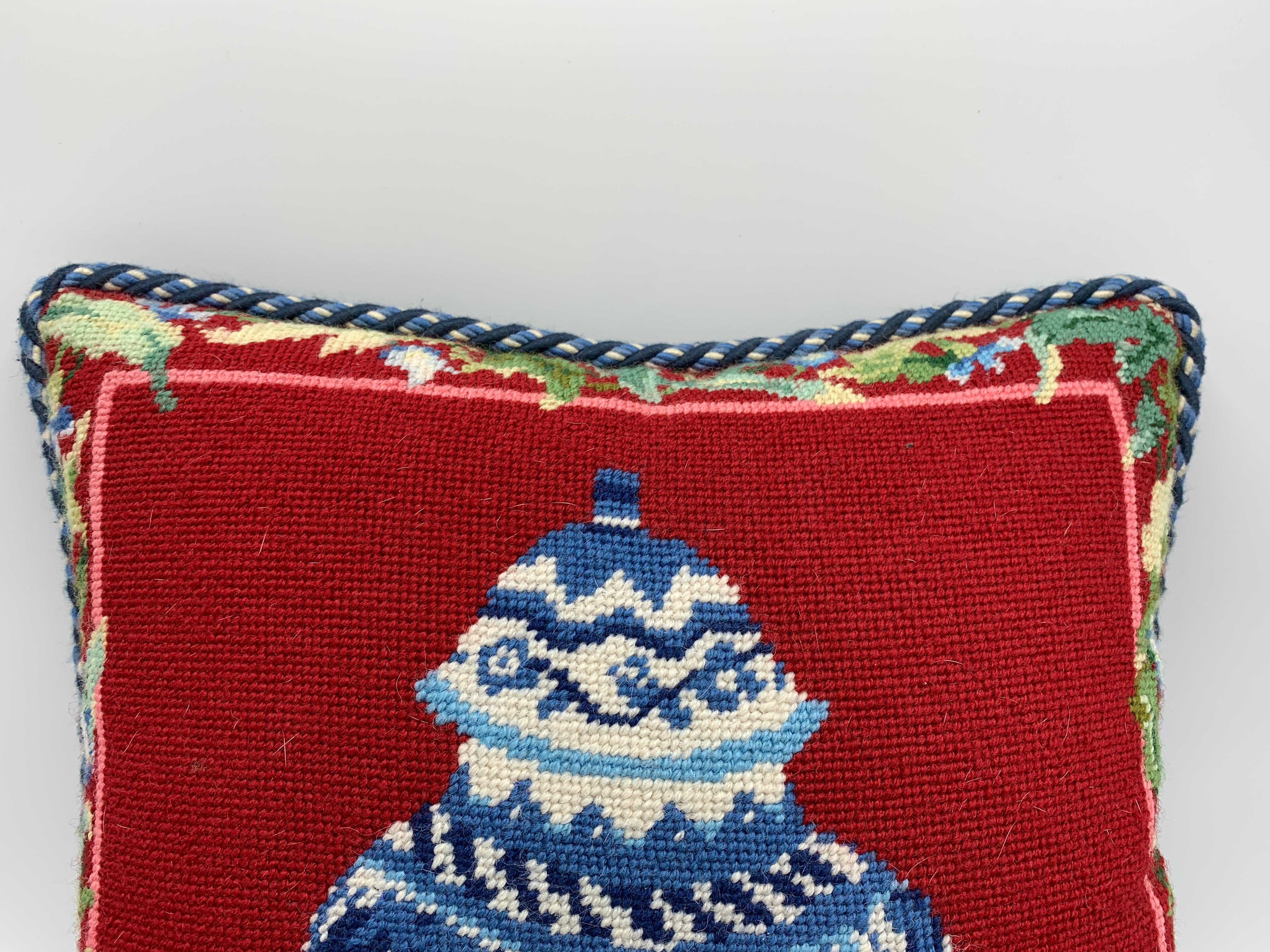 Listed is a stunning, 1970s chinoiserie needlepoint pillow. The piece has a blue and white ginger jar as the focal point, with a striking red background and floral motif along the border. Finished with a thick blue and white cord and velvet backing.