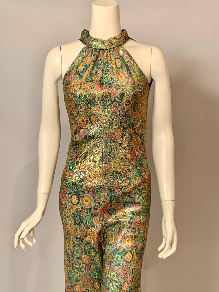 This 1970’s jumpsuit designed for Bonwit Teller is made from a gold lame fabric with floral designs in pink, turquoise, yellow, green, and lilac. The jumpsuit has a halter neckline which ties above a low cut back. The jumpsuit is fully lined in