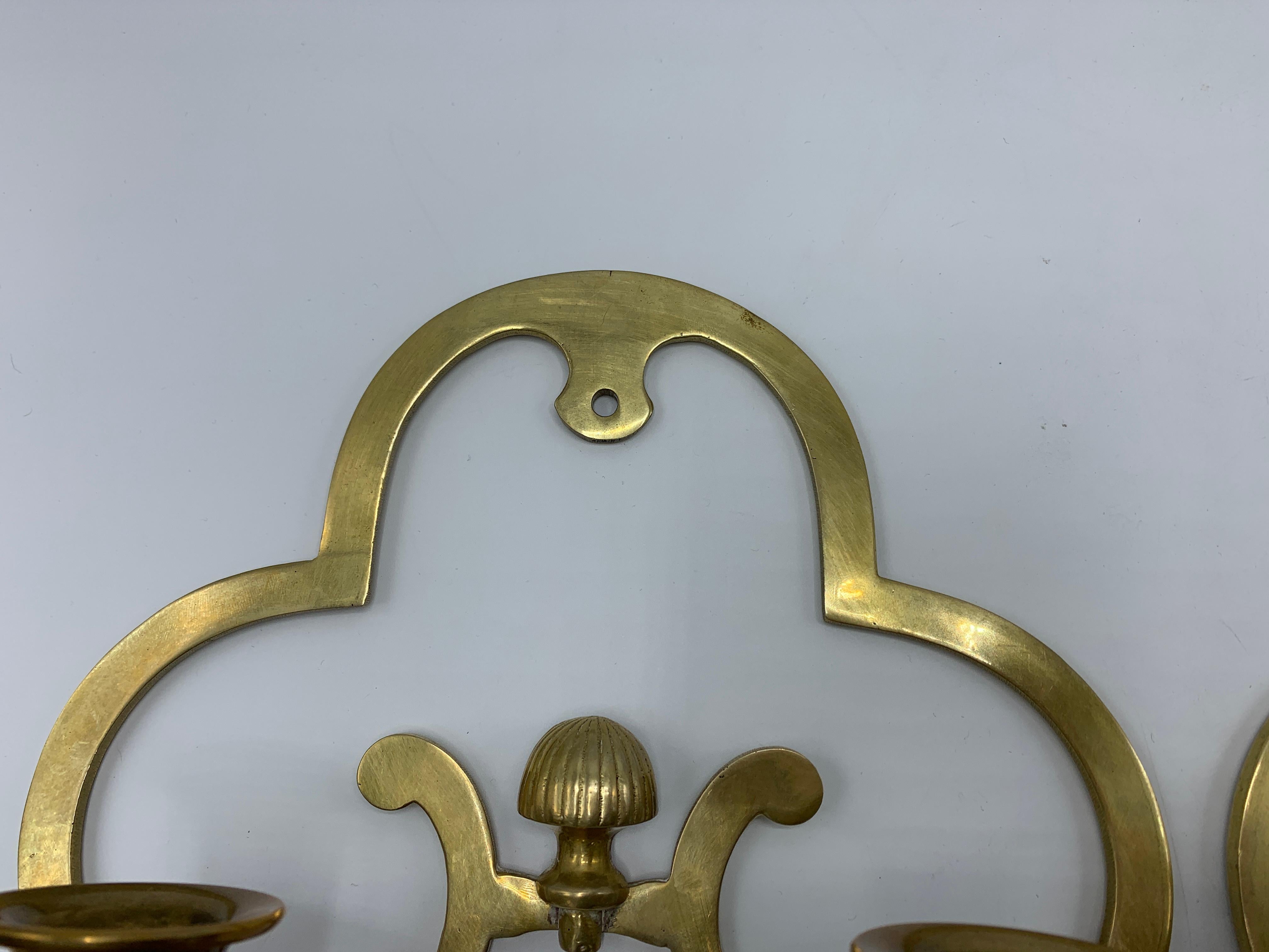 Offered is a fabulous, pair of 1970s brass candlestick wall sconces. The pair vaguely resembles the shape of a clover, with more modern curves. Hangs by nail or screw, along center.