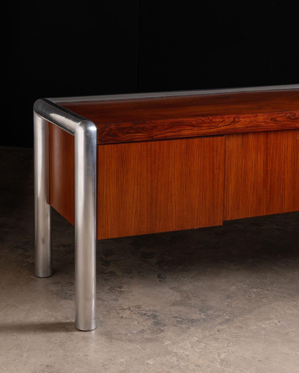 This is an exceptionally rare Brazilian Rosewood credenza designed by John Mascheroni for his TUBO series produced by Vecta Contract Furniture in the 1970s.
The satin-polished aluminum tubular frame is highly complementary to the bookmatched