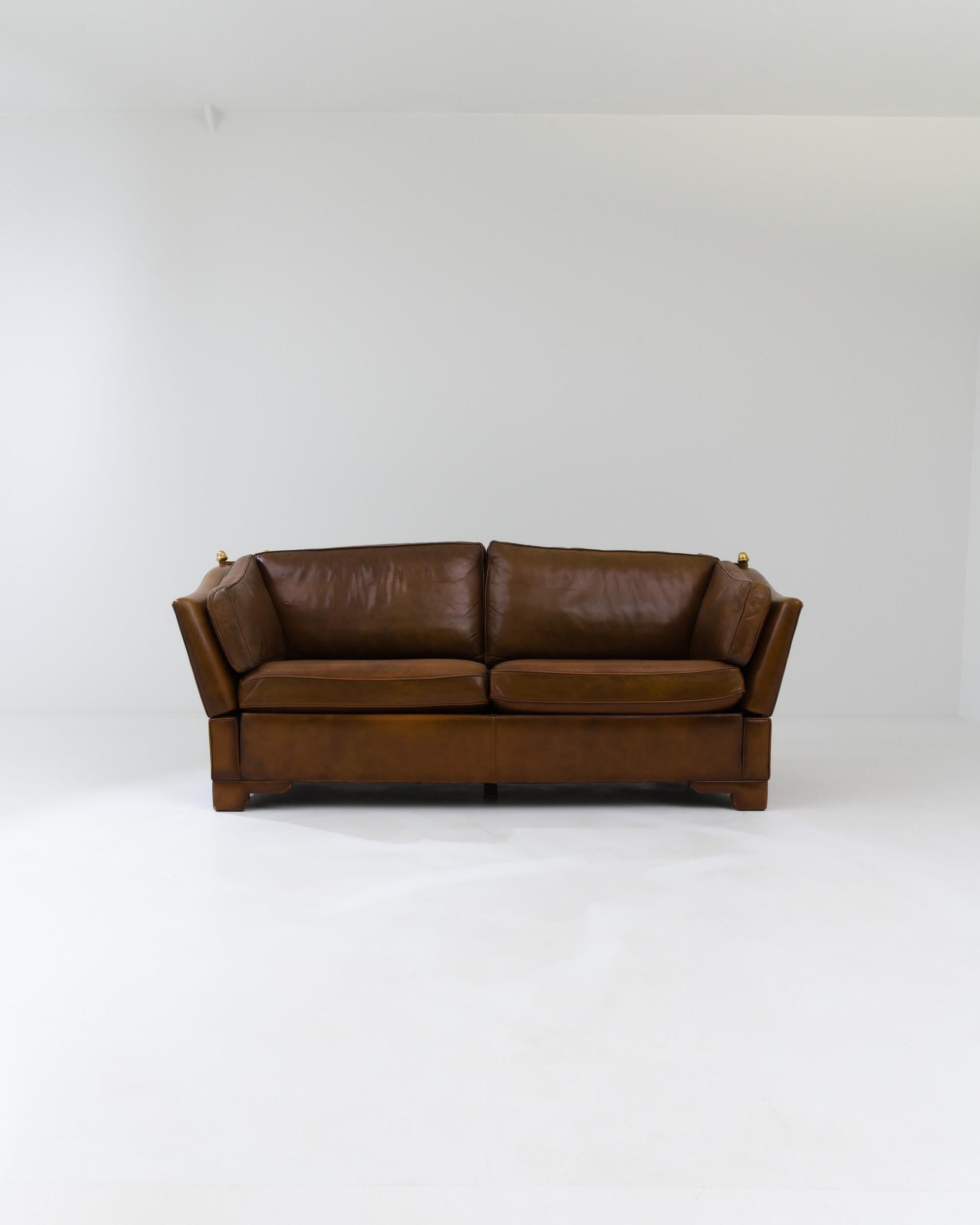 Designed in the UK circa 1970, this stylish leather two-seater sofa features an innovative design with adjustable sides that can be tightened or loosened using cords attached to the brass anchors. The tone of the high-quality leather gradually