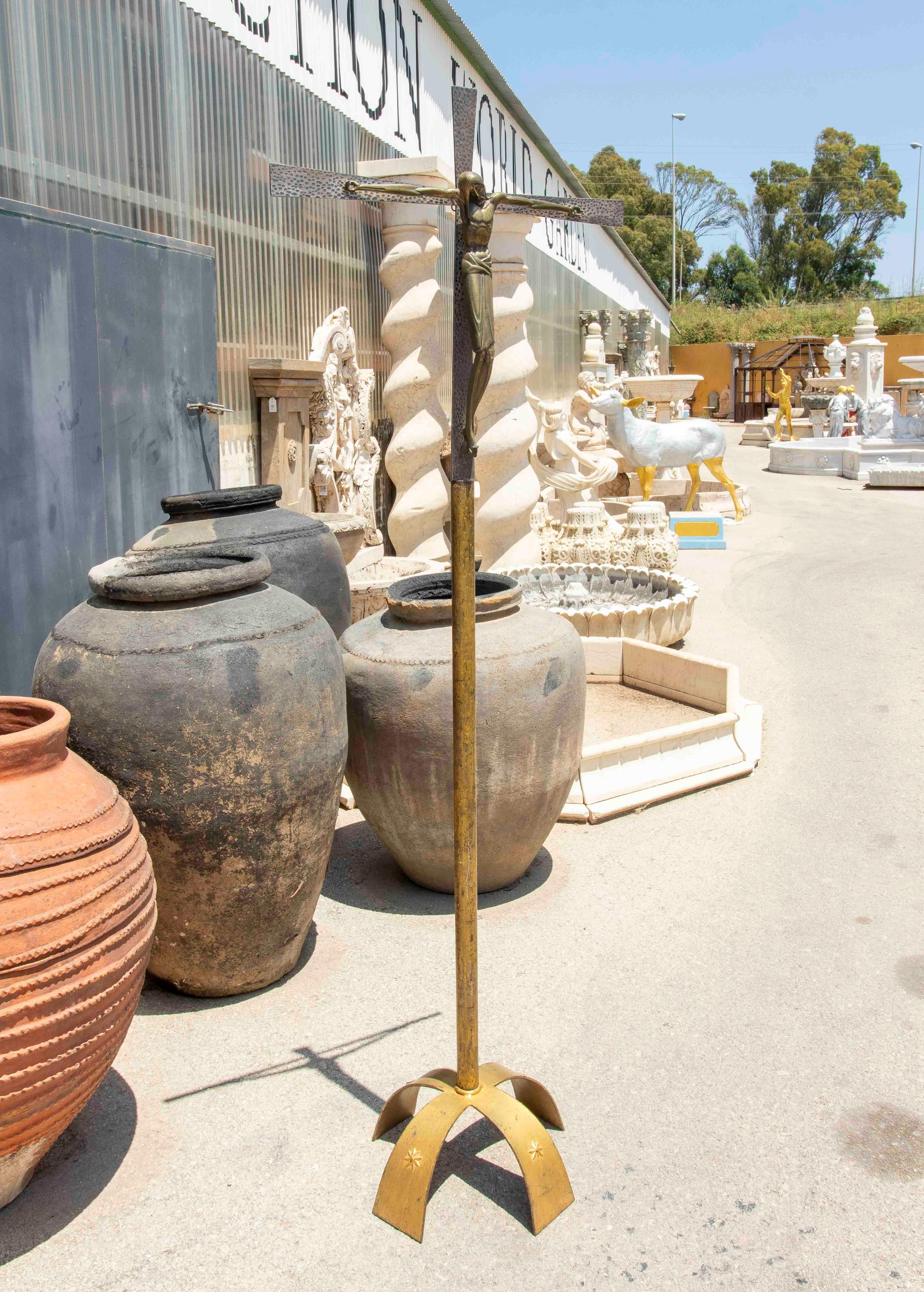 Large-format sculpture depicting Christ crucified, crafted in bronze with a hammered finish that gives it a unique, handcrafted texture. The figure of Christ is presented with outstretched arms on a stylized metal cross, supported by an elegant