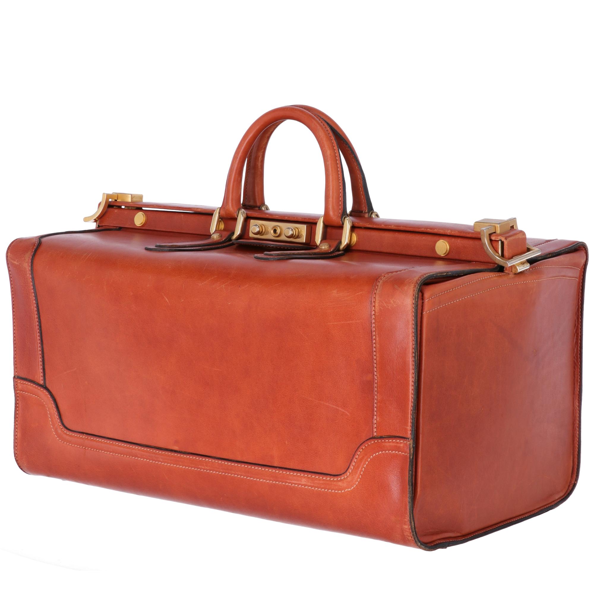 Travel trunk in brown leather, equipped with two rigid leather handles, golden metal snap closure and six feet on the bottom. Inside there are two pockets, one with zip and the other is applied closed by a leather band. Fully lined in beige