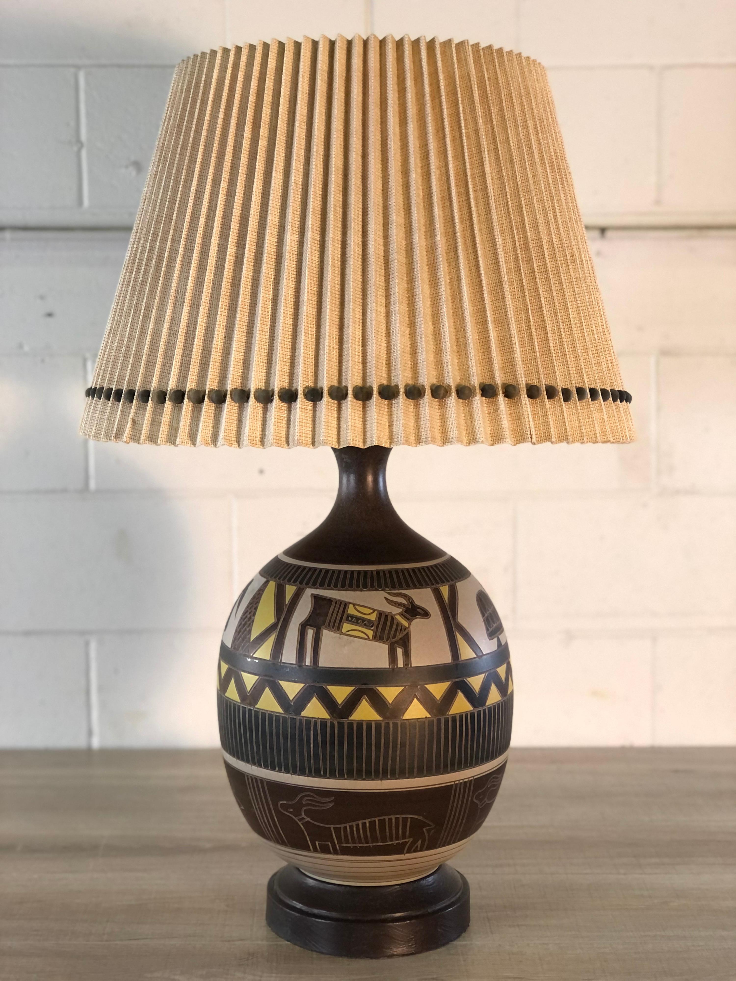 Vintage 1970s Brutalist Aztec style ceramic table lamp with the original shade. The shade has wood balls around the base of the pleated shade. The lamp has different Aztec animal scences. The lamp with out the shade measures 12” diameter x 33”
