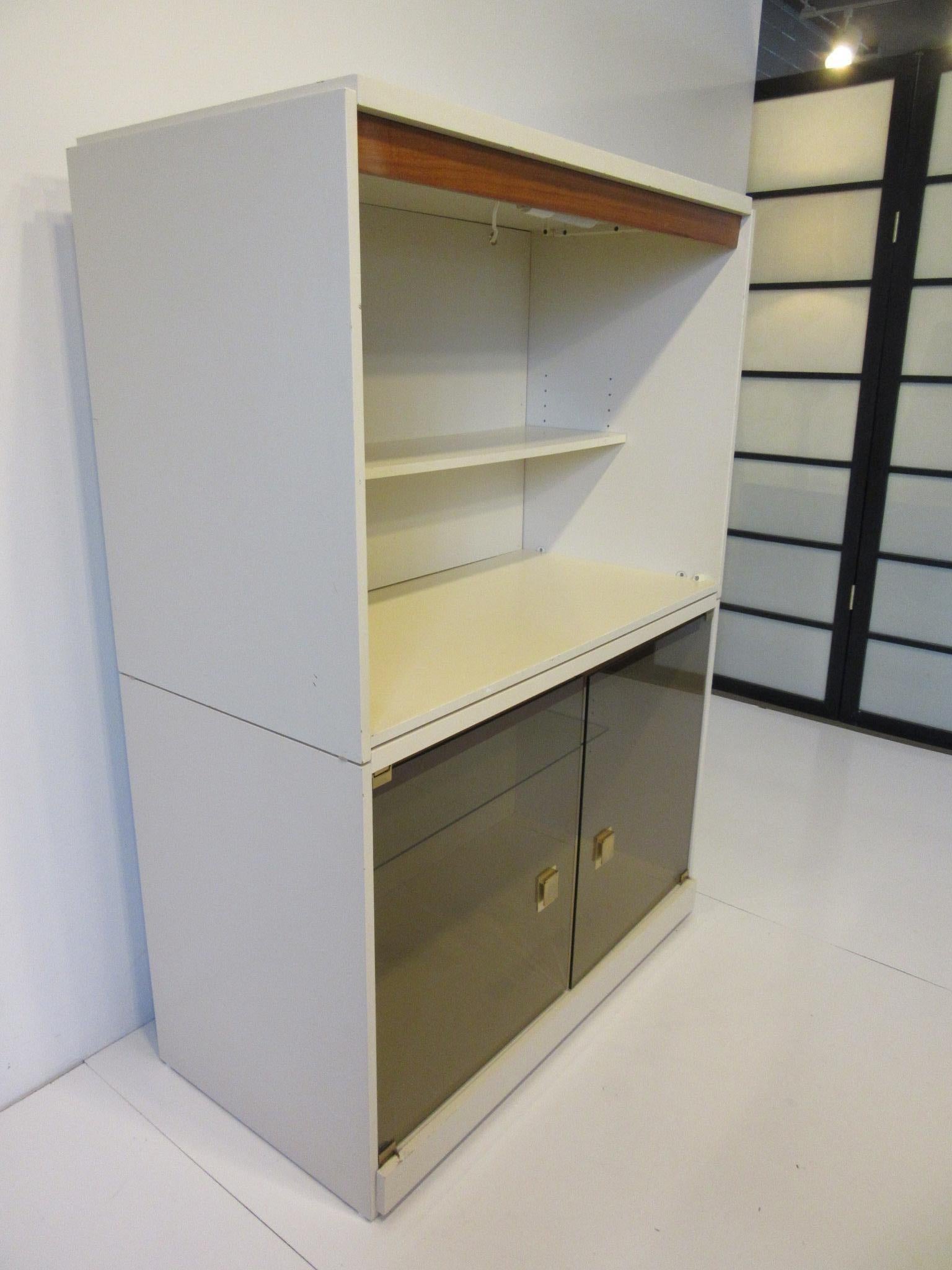 A two-piece cream lacquer finished cabinet or bookcase with adjustable shelves, smoked glass doors, brass pulls, walnut trim and lights to both units. In the manner of Milo Baughman constructed in the 1970s made in Canada.
