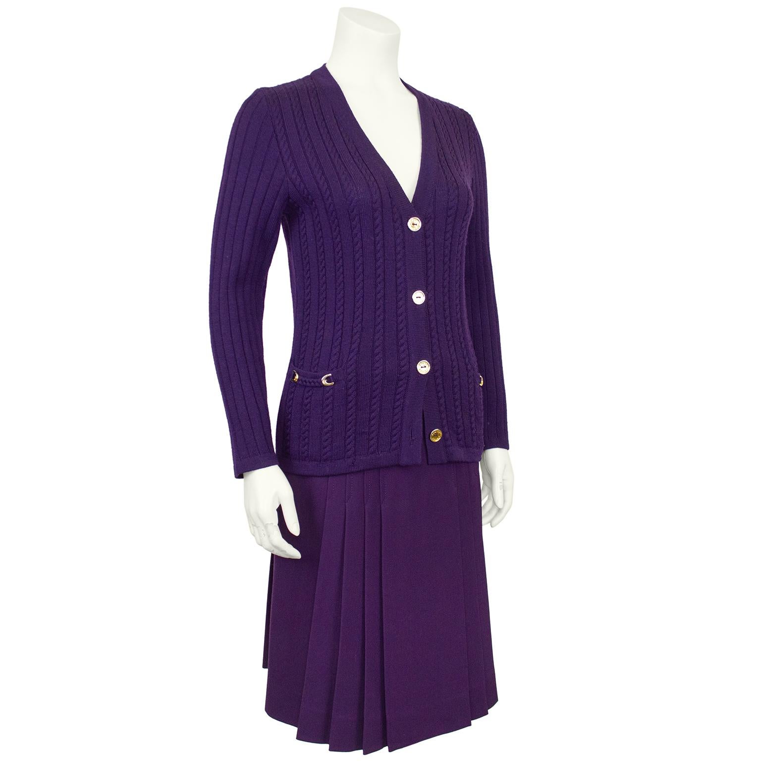 Classic and timeless Celine 100% wool jewel tone purple cardigan and gabardine skirt ensemble from the 1970's. The set features a cable knit and waffle cardigan with braided trim and gold tone metal horse shoes at the pockets. The pleated skirt is