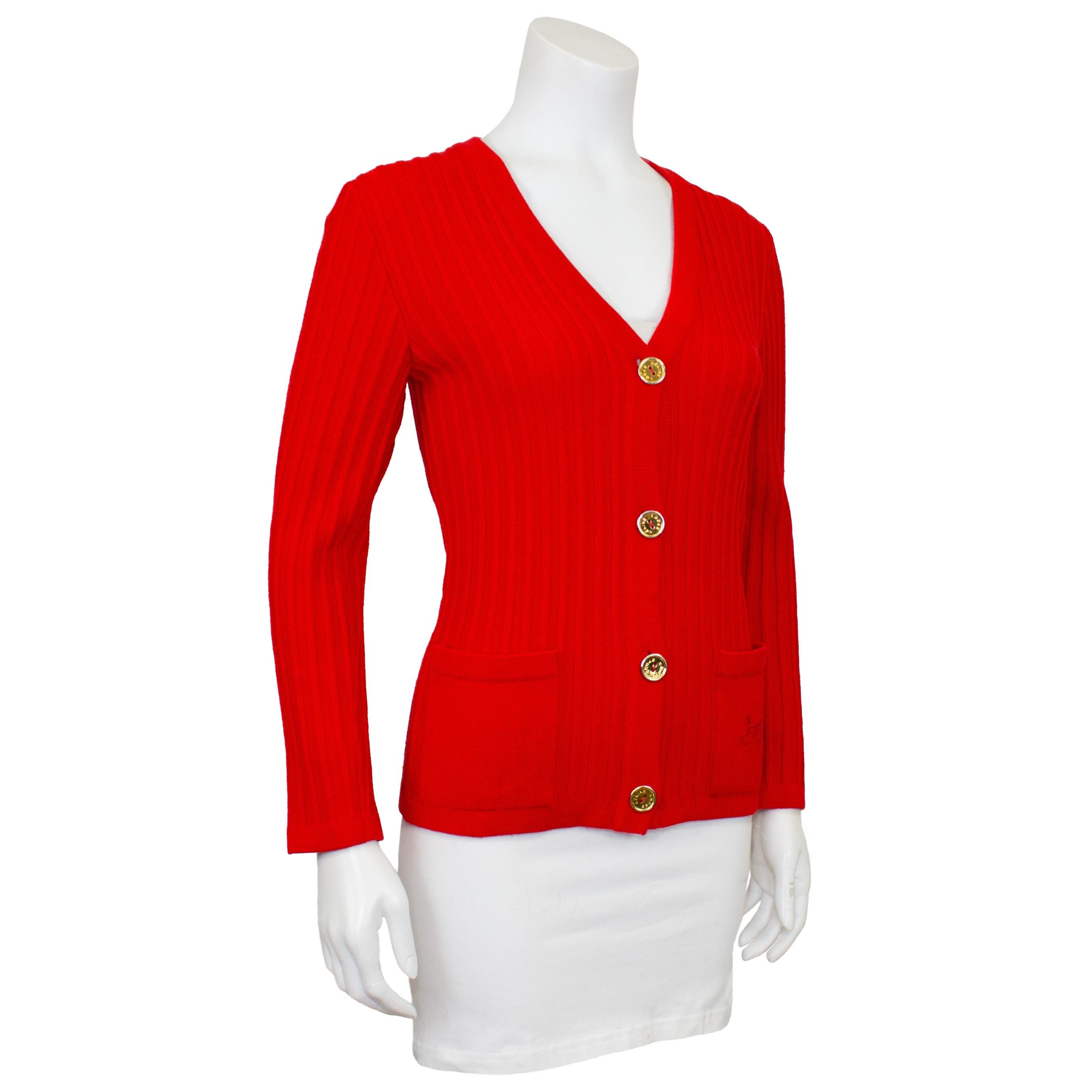 Cardigan en laine côtelée rouge vif de Celine des années 1970. Encolure en V avec de gros boutons en métal doré. Poches plaquées sur les hanches, celle de gauche (au porter) étant brodée du logo classique de Celine représentant un cheval et un