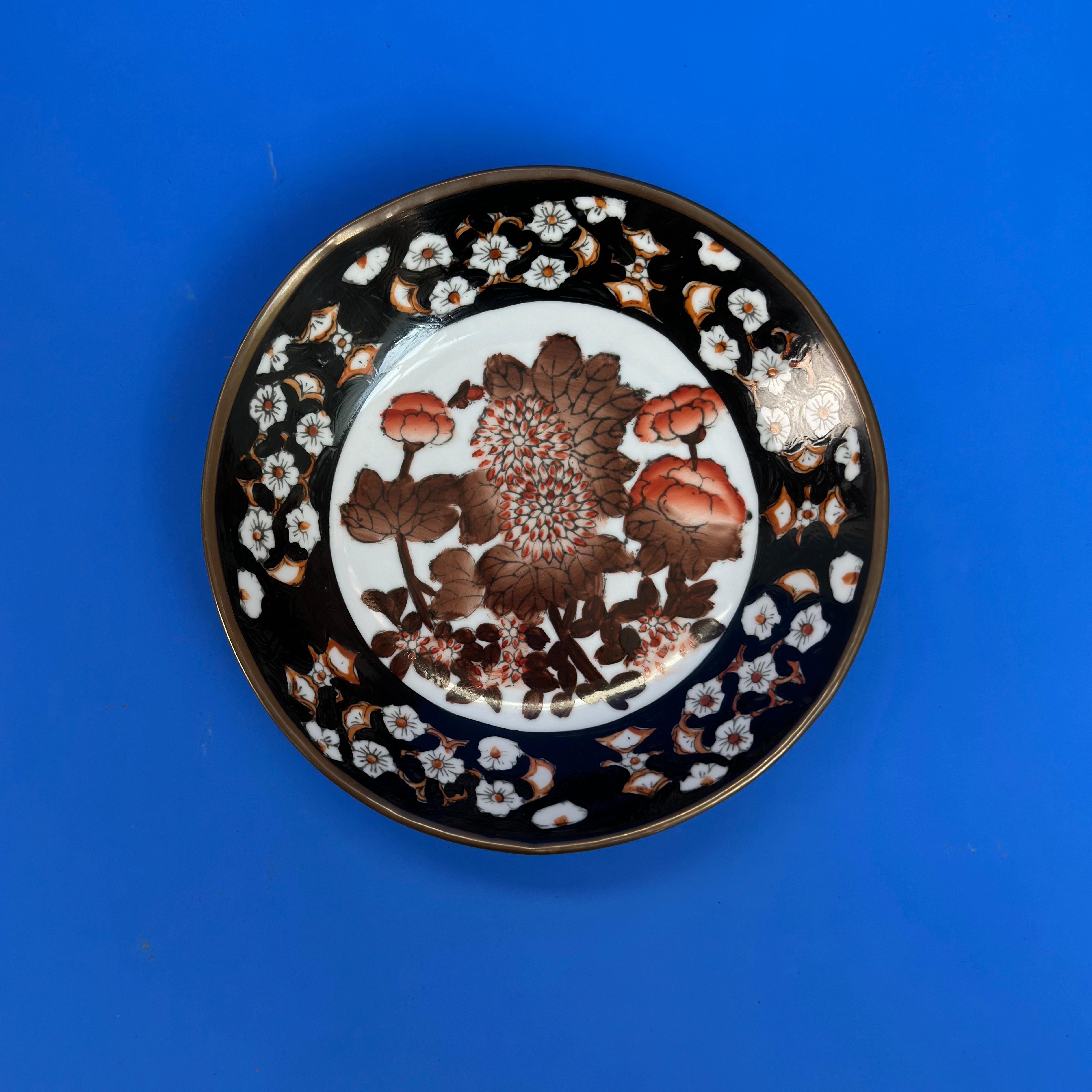 Vintage Dekorativer Wandteller

Ein japanischer Wandteller aus Vintage-Porzellan, der in den 1970er Jahren handgefertigt wurde, zeigt ein auffälliges Muster aus braunen und roten Blumen, das von einem kräftigen schwarzen Rand eingerahmt wird. Das in
