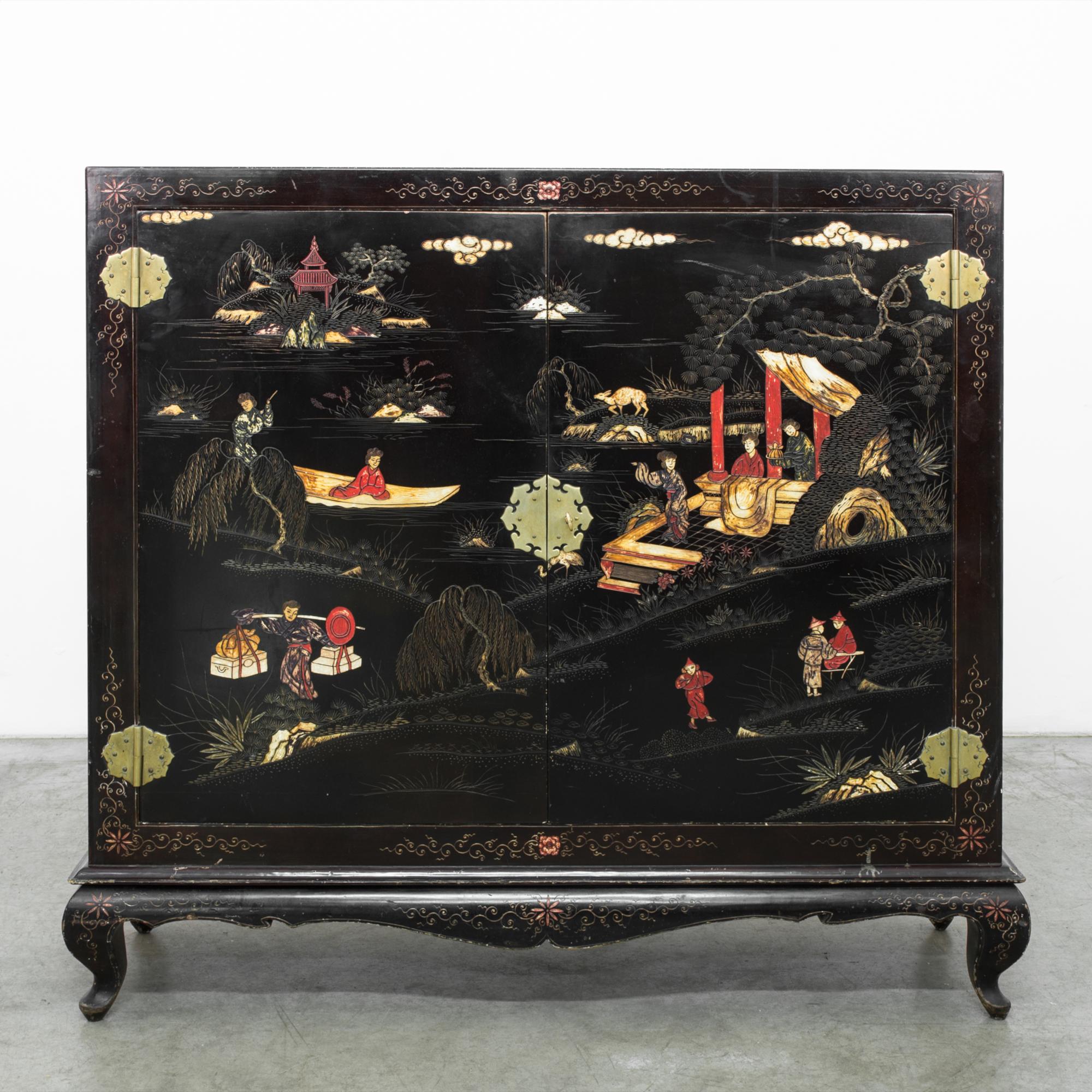 A wood patinated buffet from China, circa 1970. A beautiful black lacquered cabinet, ornately decorated with a carved and painted landscape. Double doors hold three shelves, with brass flower petal hinges and lock plate. An etched floral motif rings