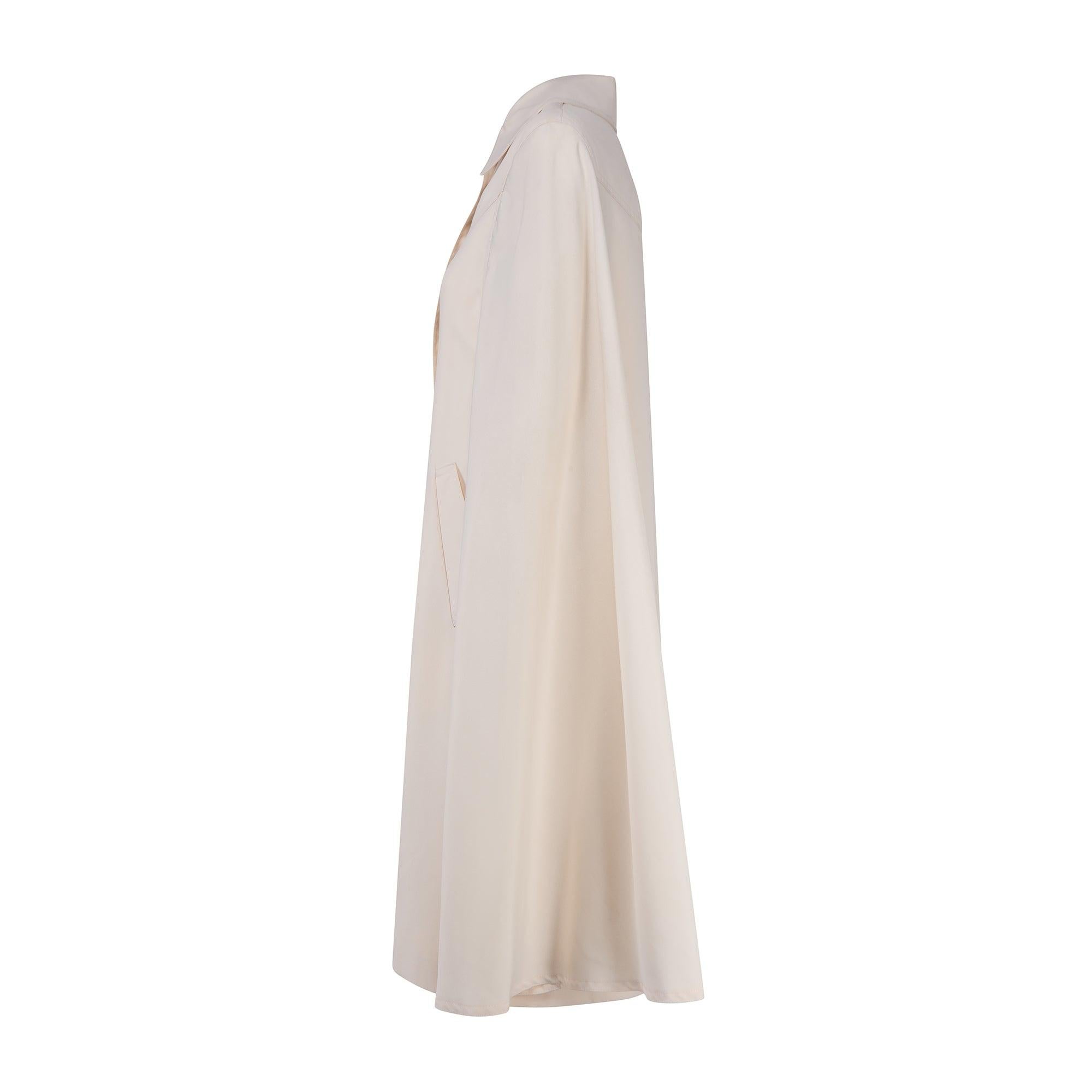 Questo grazioso midi-cape è stato realizzato negli anni '70 da un designer sconosciuto ed è tessuto in un morbido tessuto color crema. Il tessuto assomiglia a quello di un trench tradizionale e aggiunge un tocco utilitaristico a questo stile