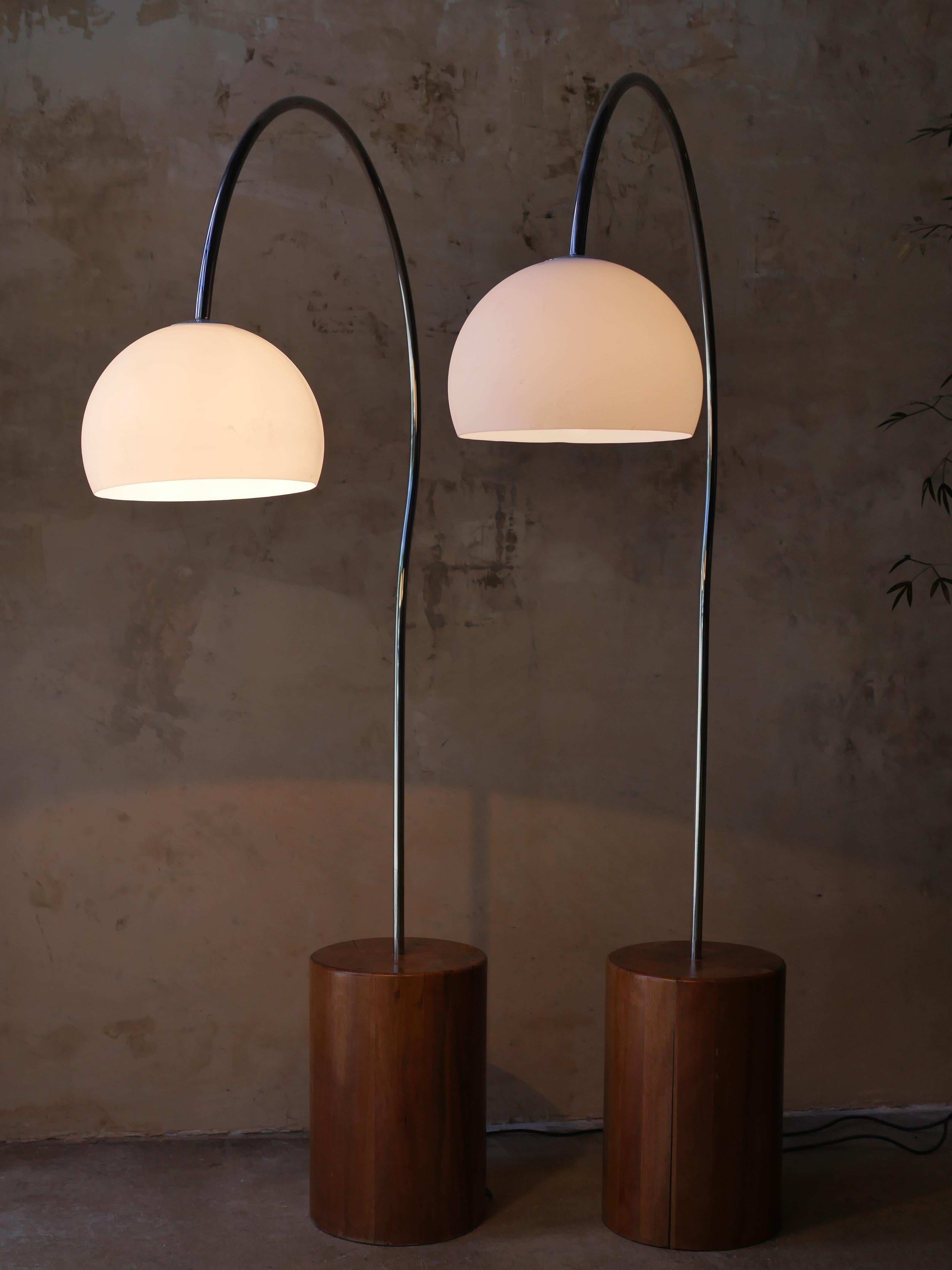 A cool Mid-Century Modern floor lamp circa 1970s, featuring a curved arm in polished chrome. Each lamp is anchored by a solid cylindrical wood base, offering a grounded contrast to the sleek metallic arcs. The opaque plastic globes cast a soft,