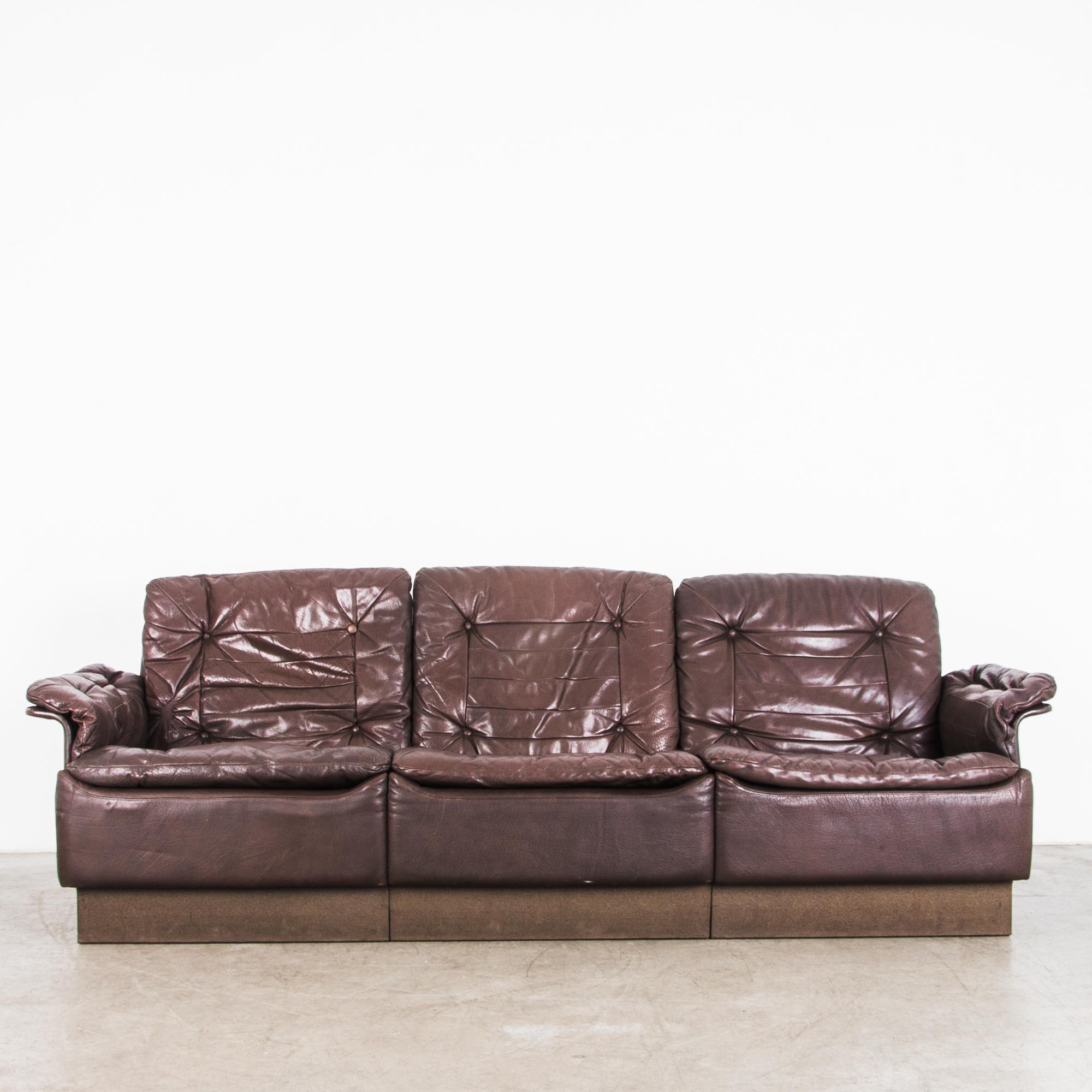 A 1970s chocolate brown leather sofa, built in Denmark. The buttoned sofa cushions and arm-rests speak to classic arm-chair design, while clean curves and elevating platform provide a modernist silhouette. The rich color, fine quality of the leather