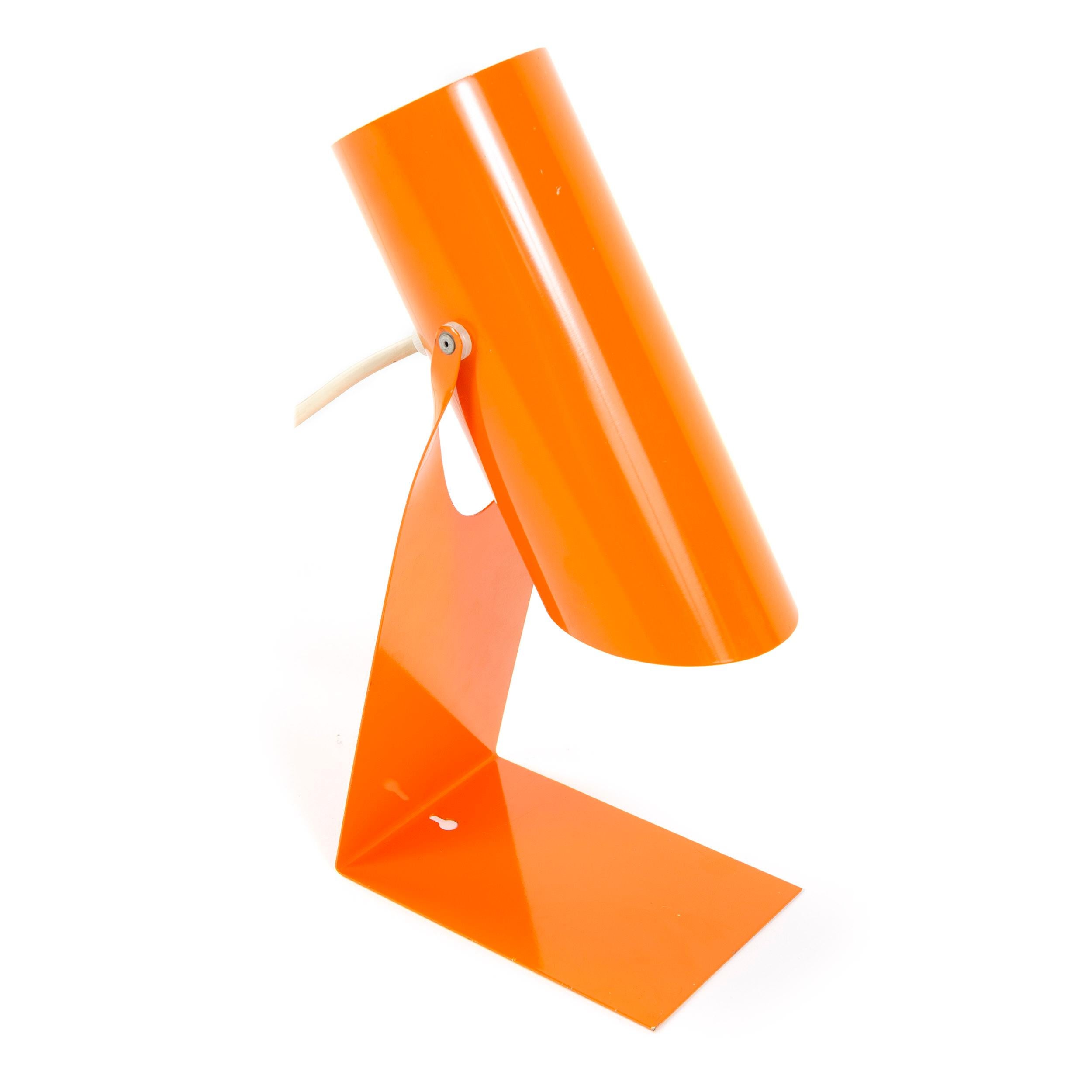 A versatile and vibrant orange enamel lighting device that primarily functions in a desk or table capacity but has a keyhole slot which allows it to function additionally as a wall lamp. The base is one piece of flat, folded steel cradling a