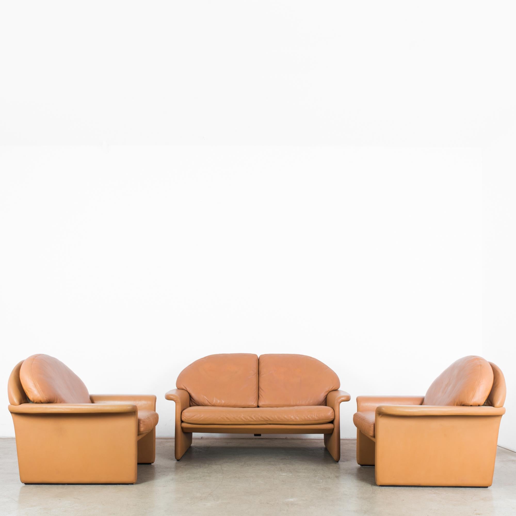 A 1970s leather sofa by Swiss furniture manufacturers De Sede, accompanied by a pair of matching armchairs. A light, square frame of camel colored leather, vaulted by the cushioned arches of the backrests. The armrests turn outward with a coy flick.