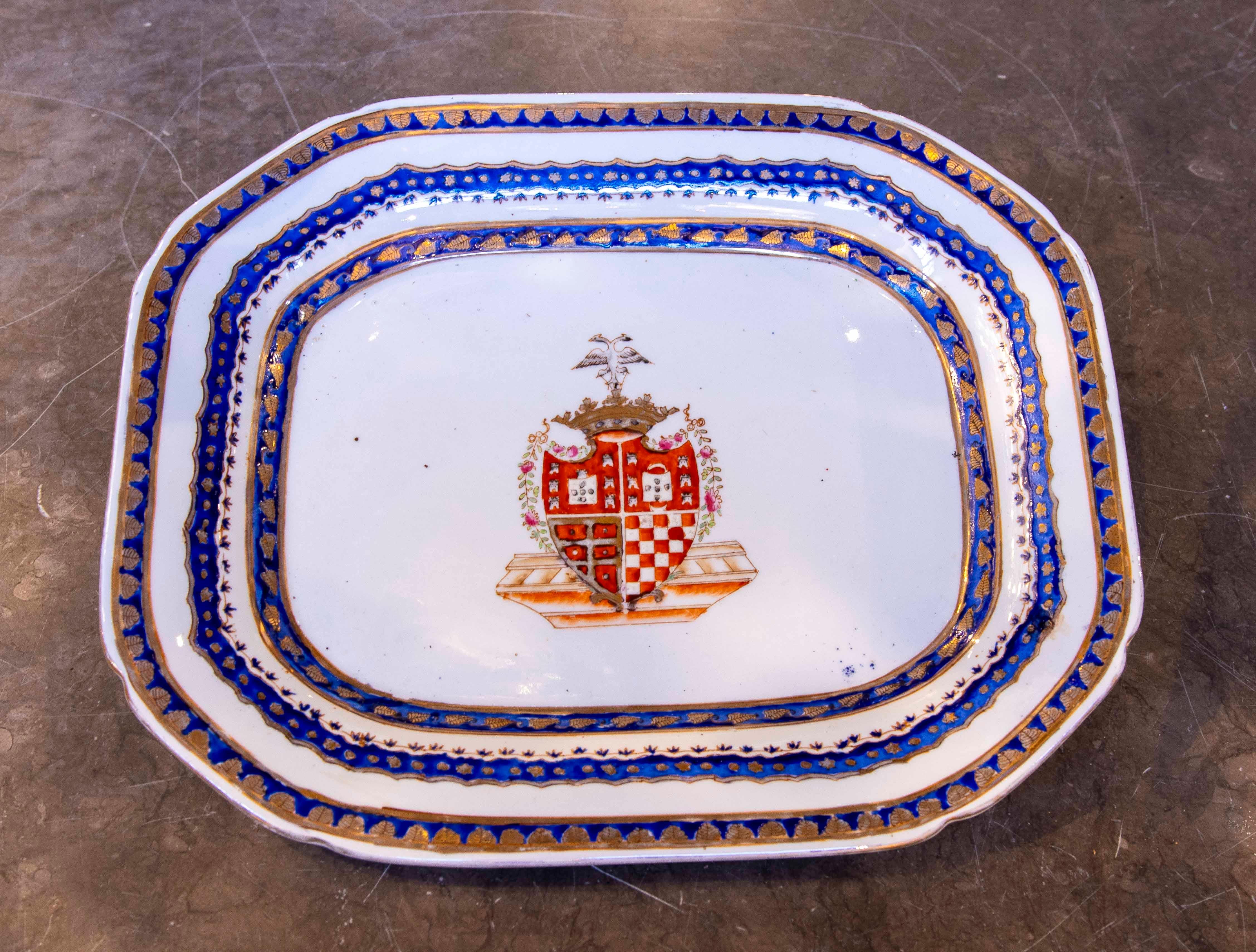 Assiette de reproduction décorative en céramique fine, au design rectangulaire et aux bords biseautés, ornée de motifs dorés et bleus encadrant un blason central. Le blason présente des détails héraldiques sur une base aux tons chauds. Au bas de la