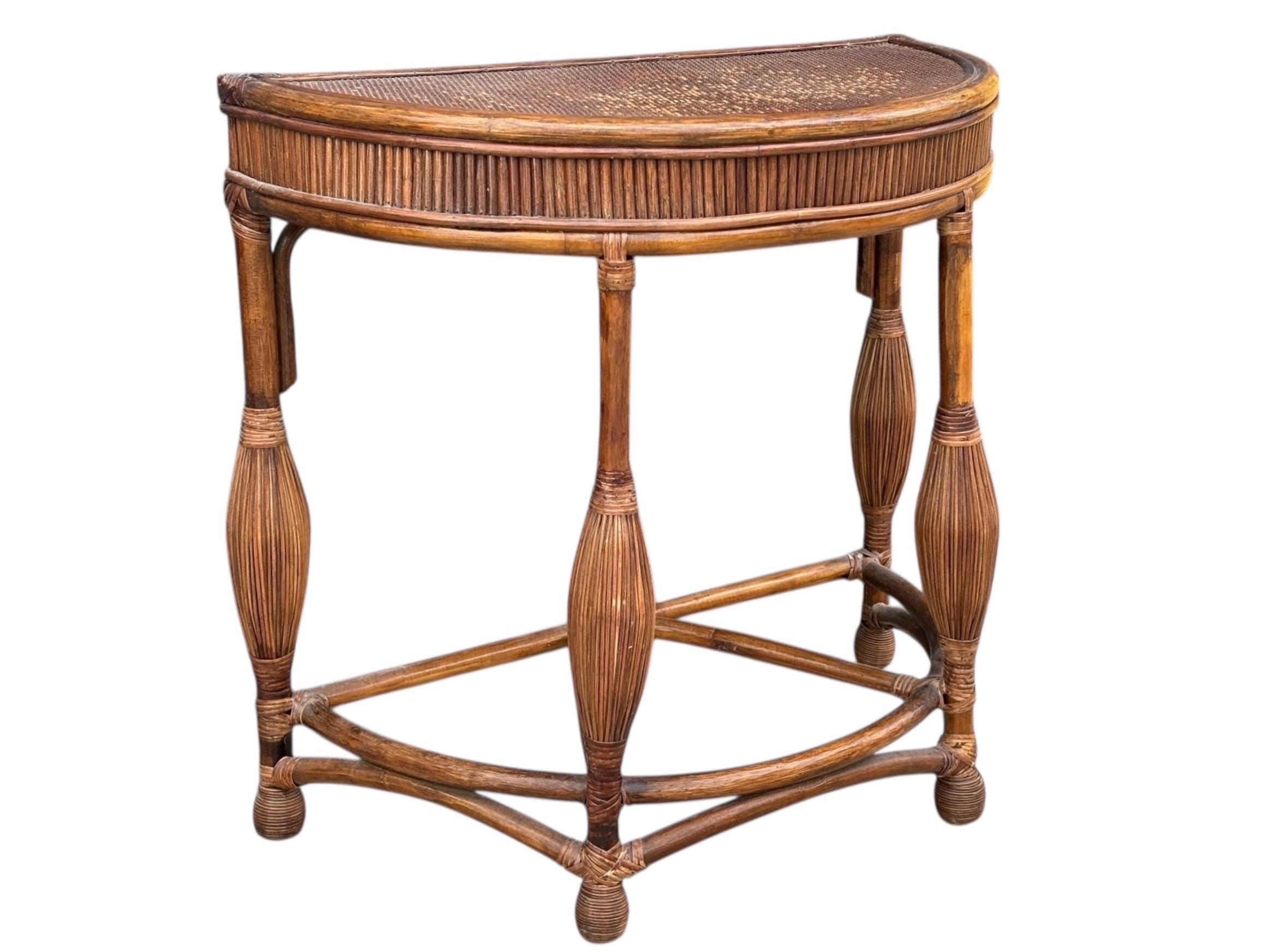 1970's demilune hall table. A bamboo frame is secured on the joins with rattan wrapping. The demilune table top is covered with woven rattan, and decorated with a pencil wood table skirt. Legs decorated with rattan balusters are supported with a