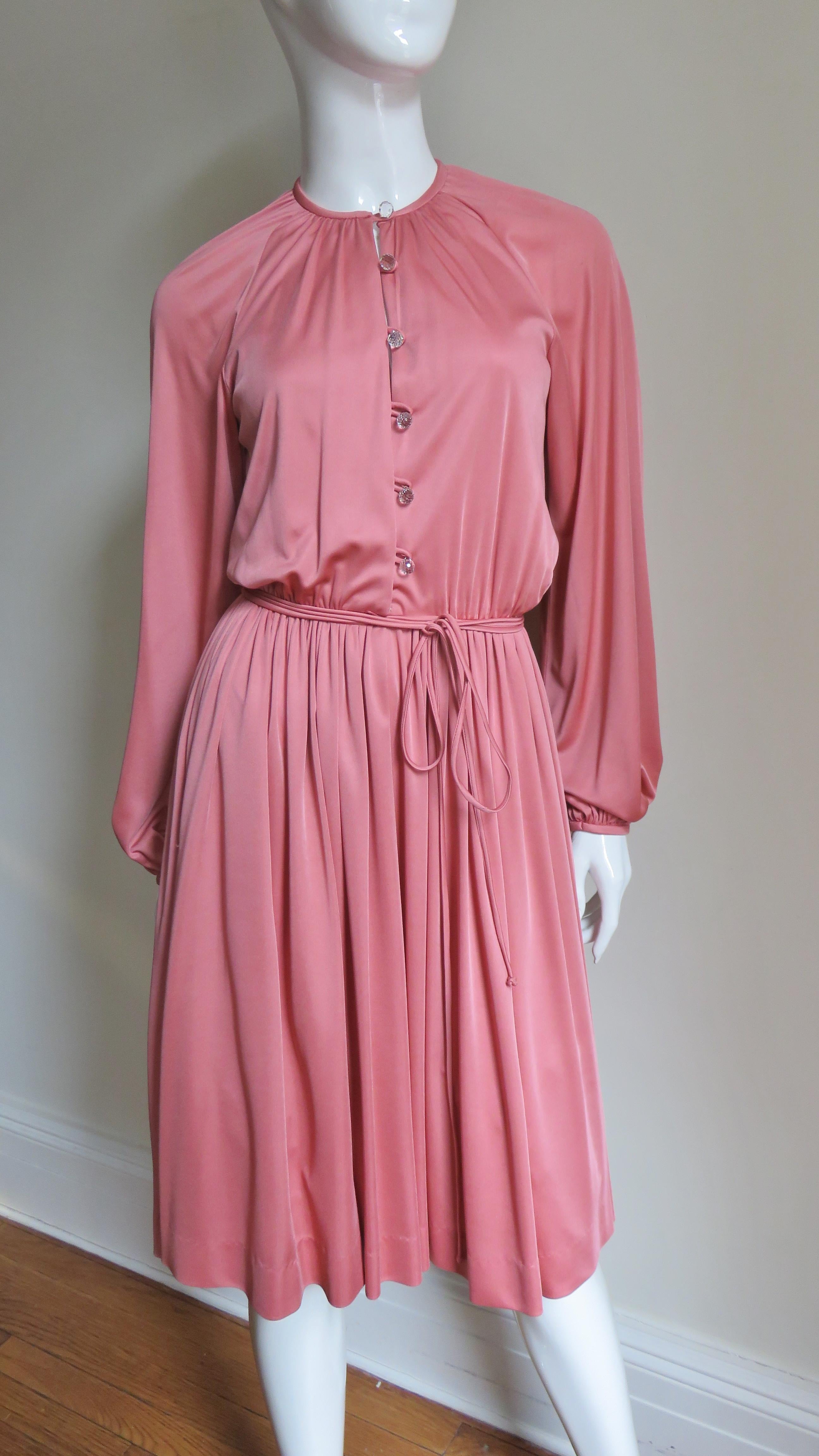 A pretty pink jersey dress by Donald Brooks. It has crew neckline and raglan balloon sleeves.  The bodice has beautiful round faceted cut crystal buttons and loops up the front.  The skirt gathers onto the bodice which has a small piped tie belt at