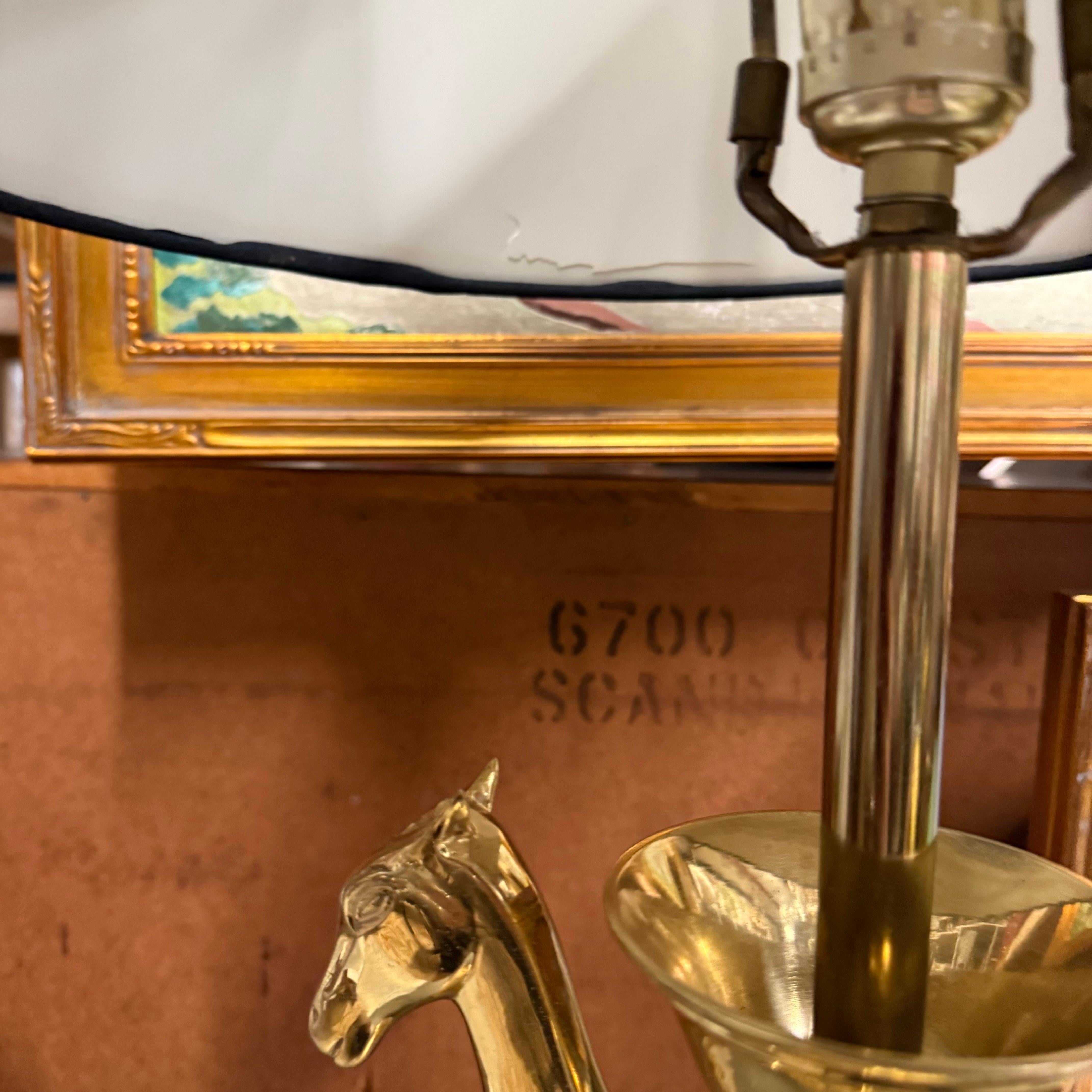 This fantastic lamp features two symmetrical horse head figures in brass.  The sculptural detailing includes stylized, elongated horse necks that support a central bowl-like structure. It rests on a tiered, circular base with curved feet. The black