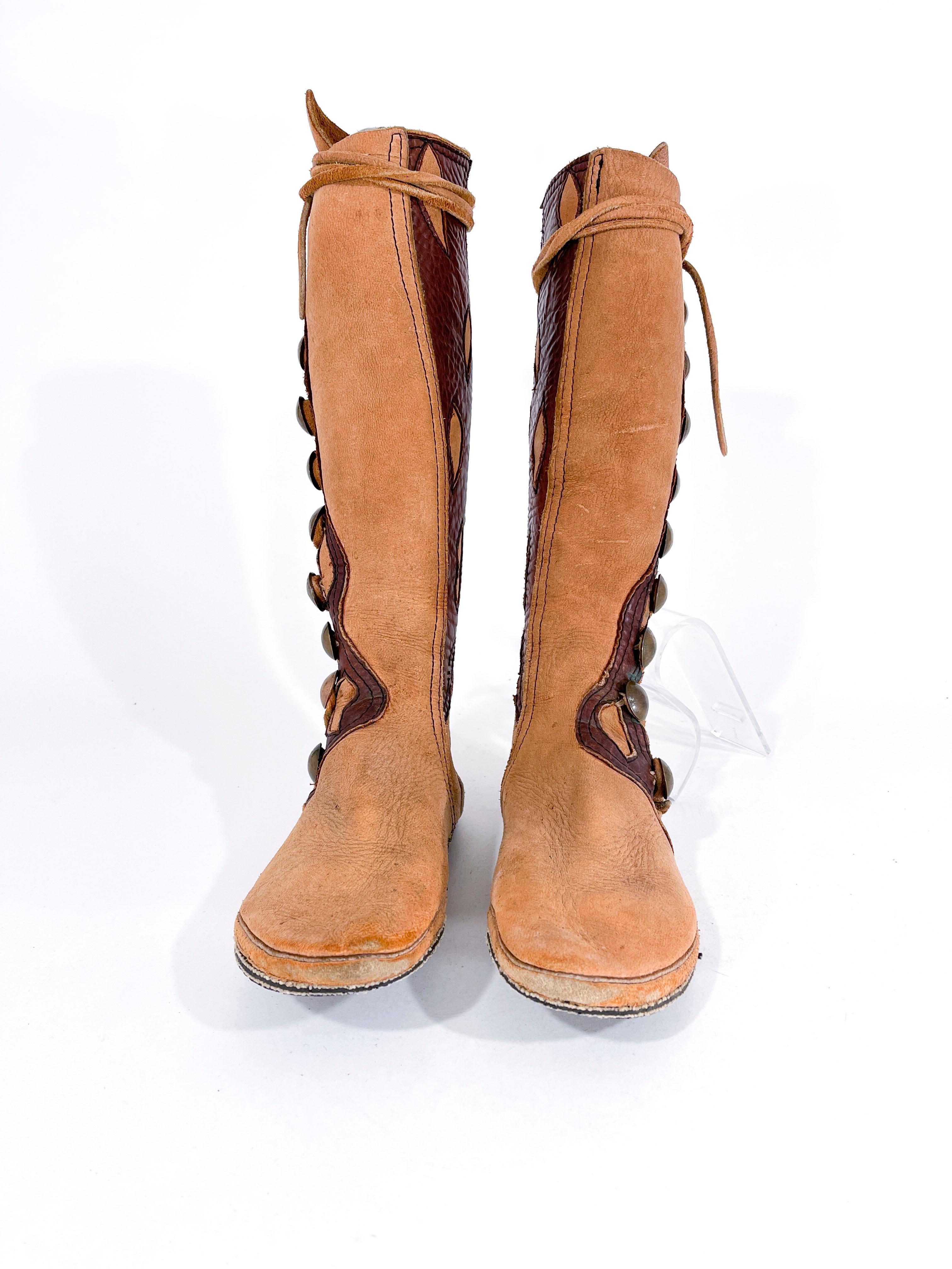 1970s custom-made elk hide bohemian gladiator style boots featuring contrasting leather inserted detailing and copper coin decorative buttons. The boot is knee-high length. 