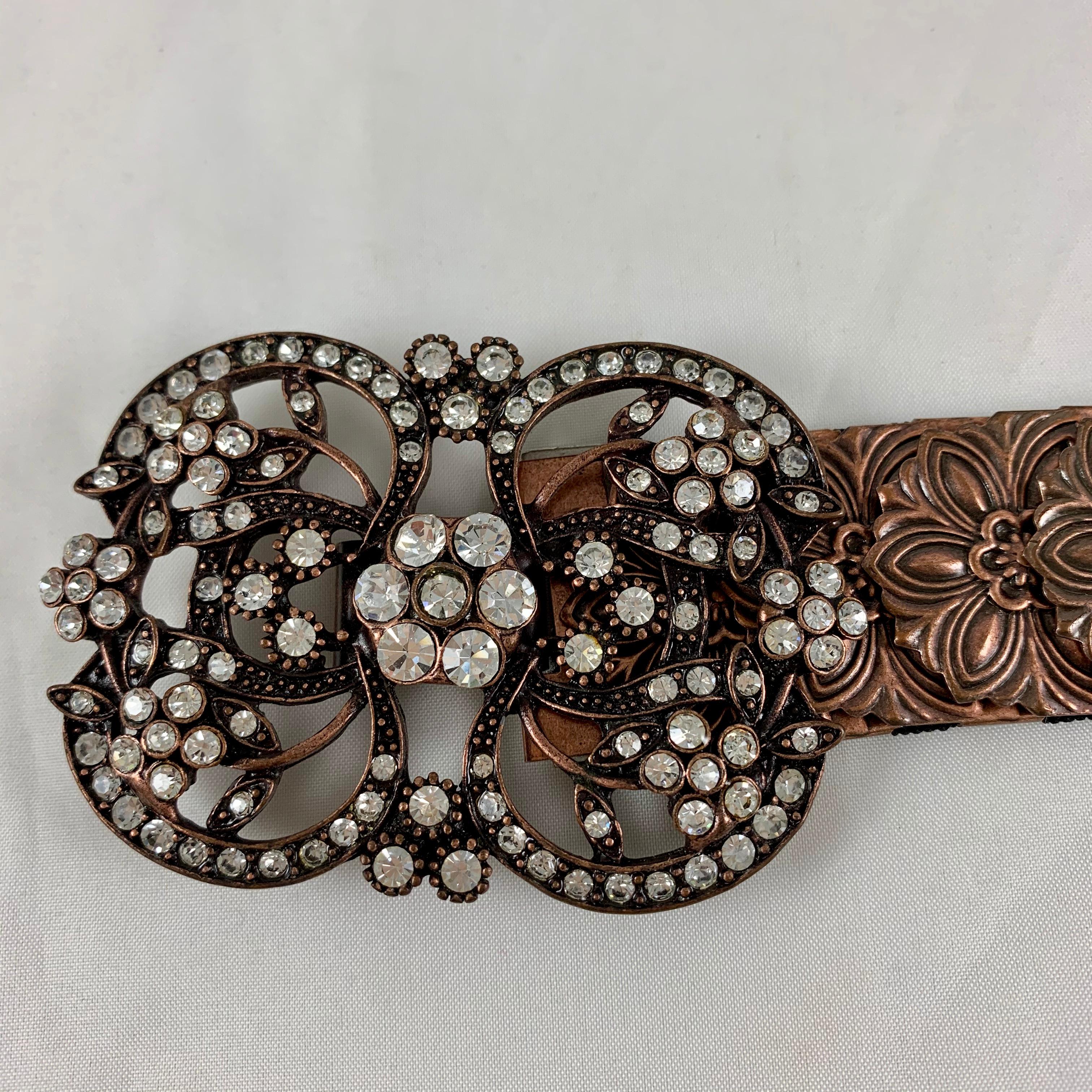 A 1970s era belt made of copper-tone metal snake scale links with a metal and crystal jeweled medallion buckle. Very well made.

Assembled by hand, the links show a stamped floral pattern attached to a black elasticized fabric backing. The links