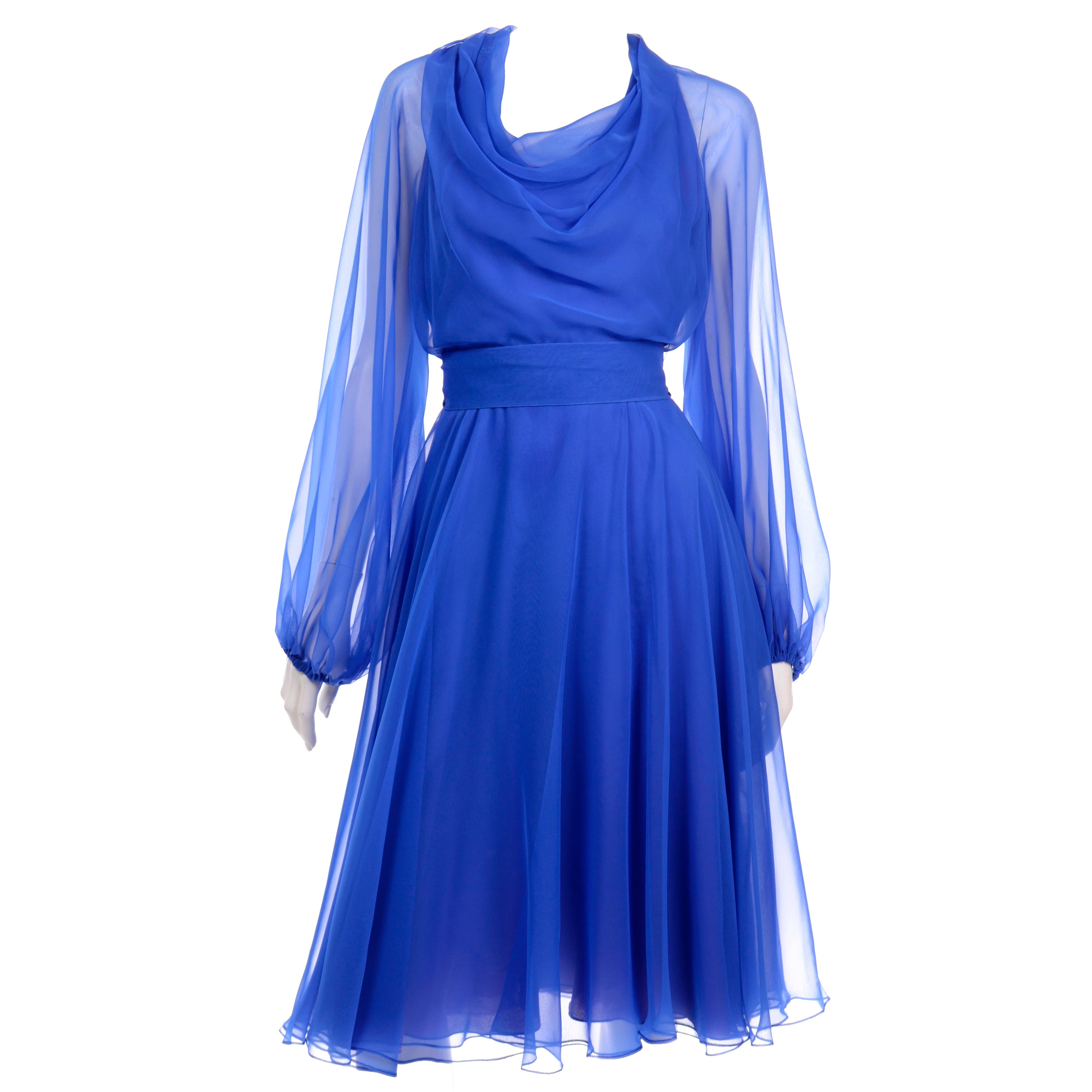 This is a 1970's vintage blue chiffon dress from Estevez. The dress has sheer sleeves, a shawl neckline and a full skirt.  There is a fabric sash and the dress closes with a back zipper. The dress has blousy sleeves with elastic cuffs and the waist