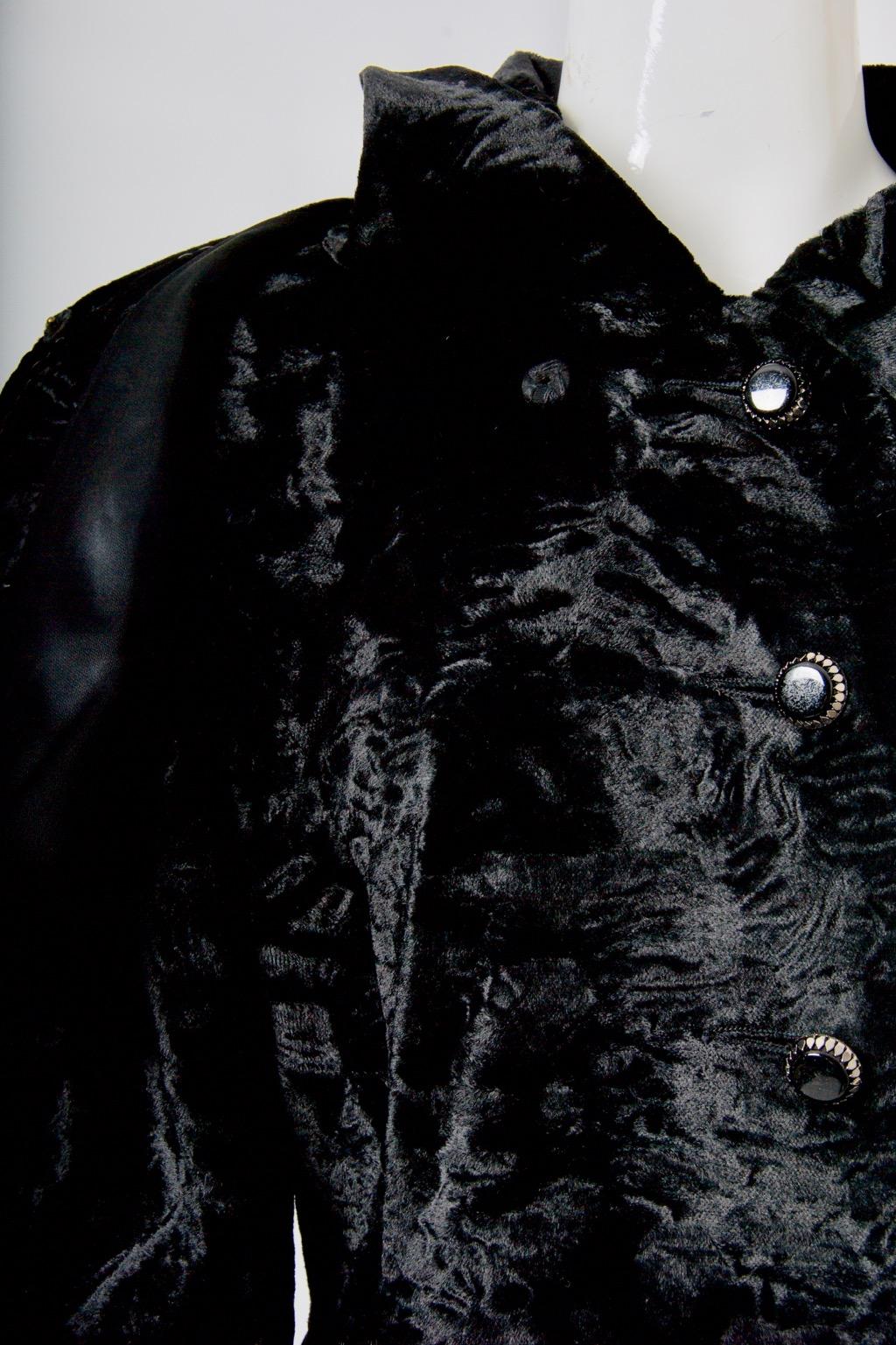 Schwarzer Mantel aus Astrachan-Kunststoff mit abnehmbarem Umhang. Der taillierte, prinzessinnenhafte Body ist einreihig und wird mit 8 schwarzen Stein- und Silberknöpfen bis zum Halsausschnitt geschlossen; seitliche Schrägtaschen. Schwarzes Futter.