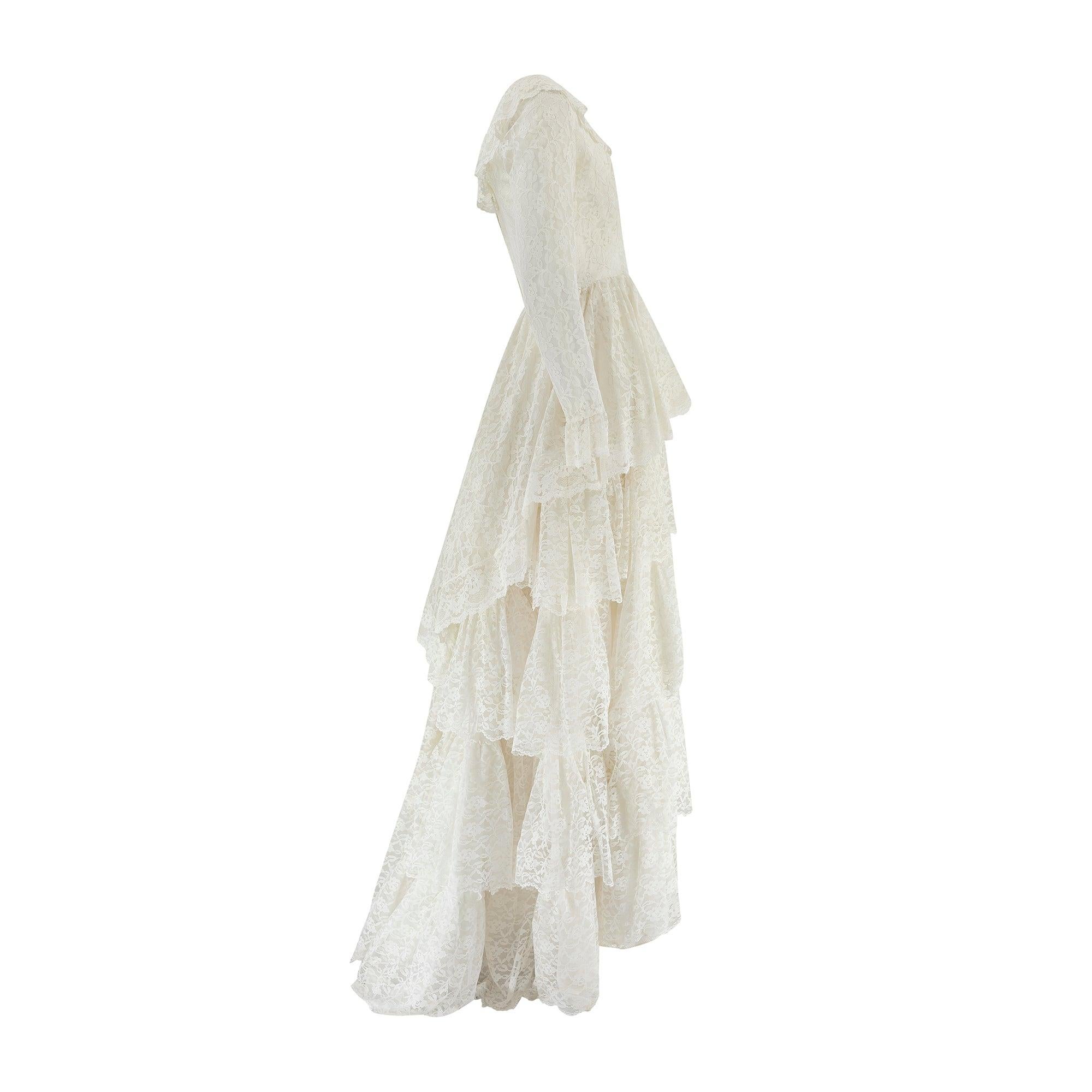 Made in the USA circa 1975, this enchanting wedding dress, with its tiered construction and layers of delicate lace, is an unforgettable choice for any vintage-loving bride. It has a long-sleeved top with a fitted bust and wide, rounded neckline